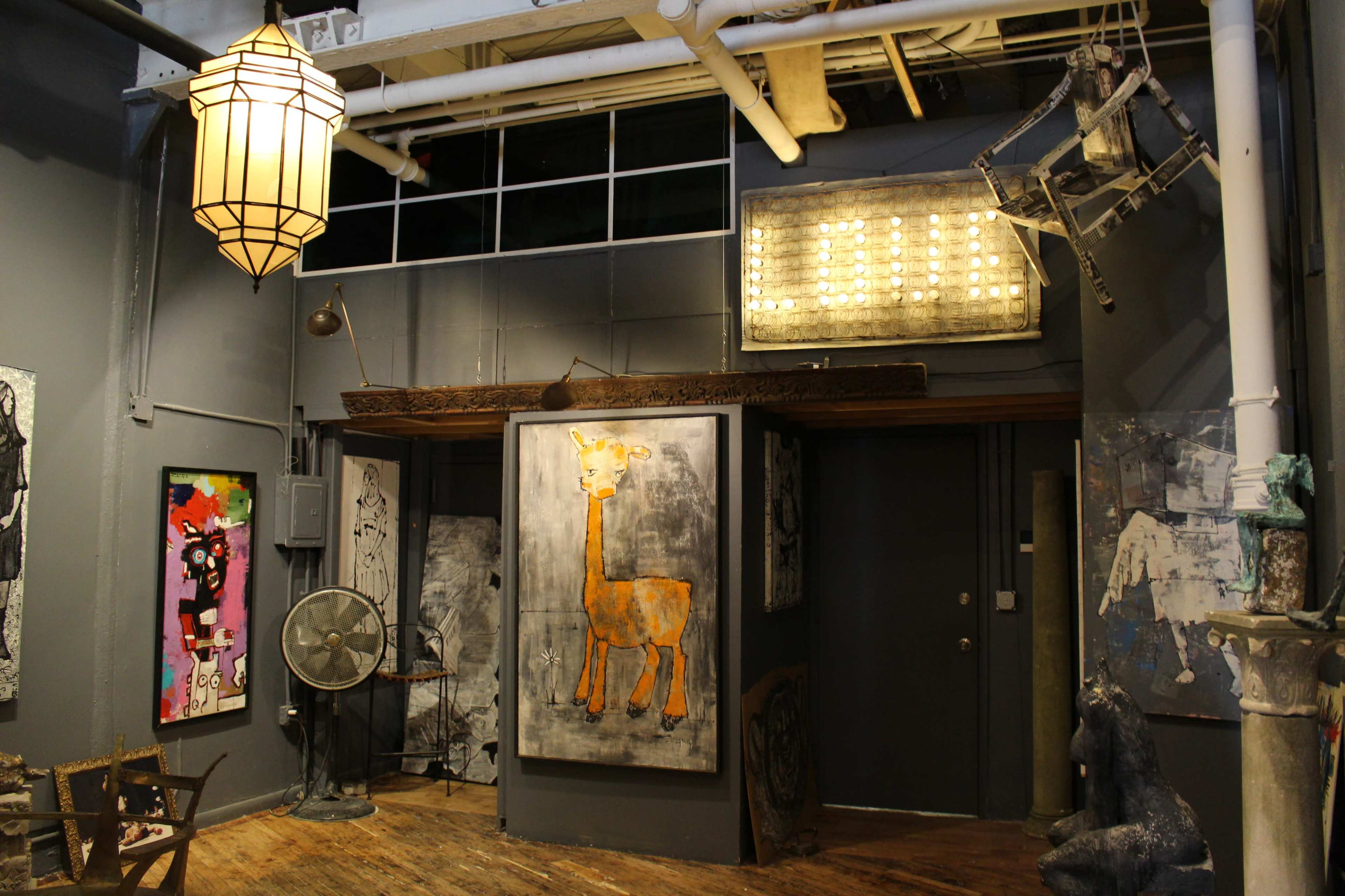 The image depicts an art-filled interior space featuring various artworks, including a painting of a giraffe, alongside sculptures and a large illuminated sign.