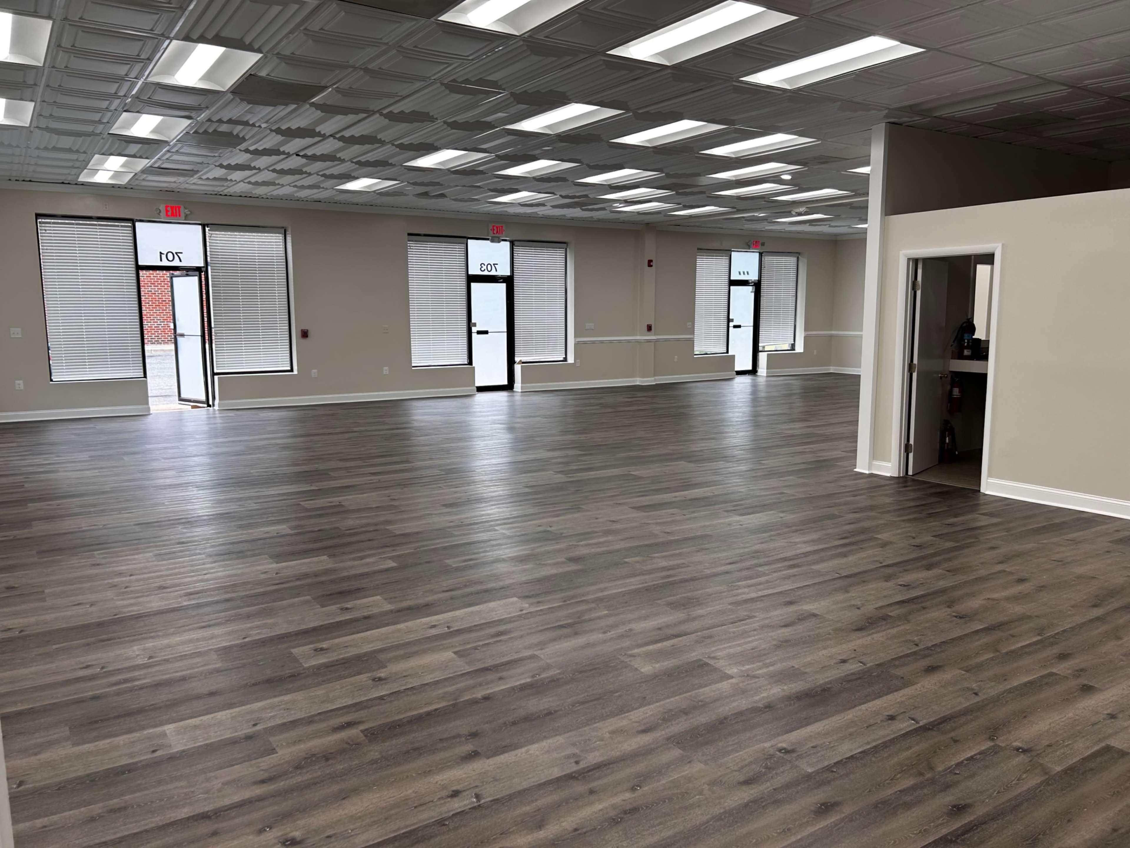 The image shows a spacious, empty room with wooden flooring and multiple windows along the walls.