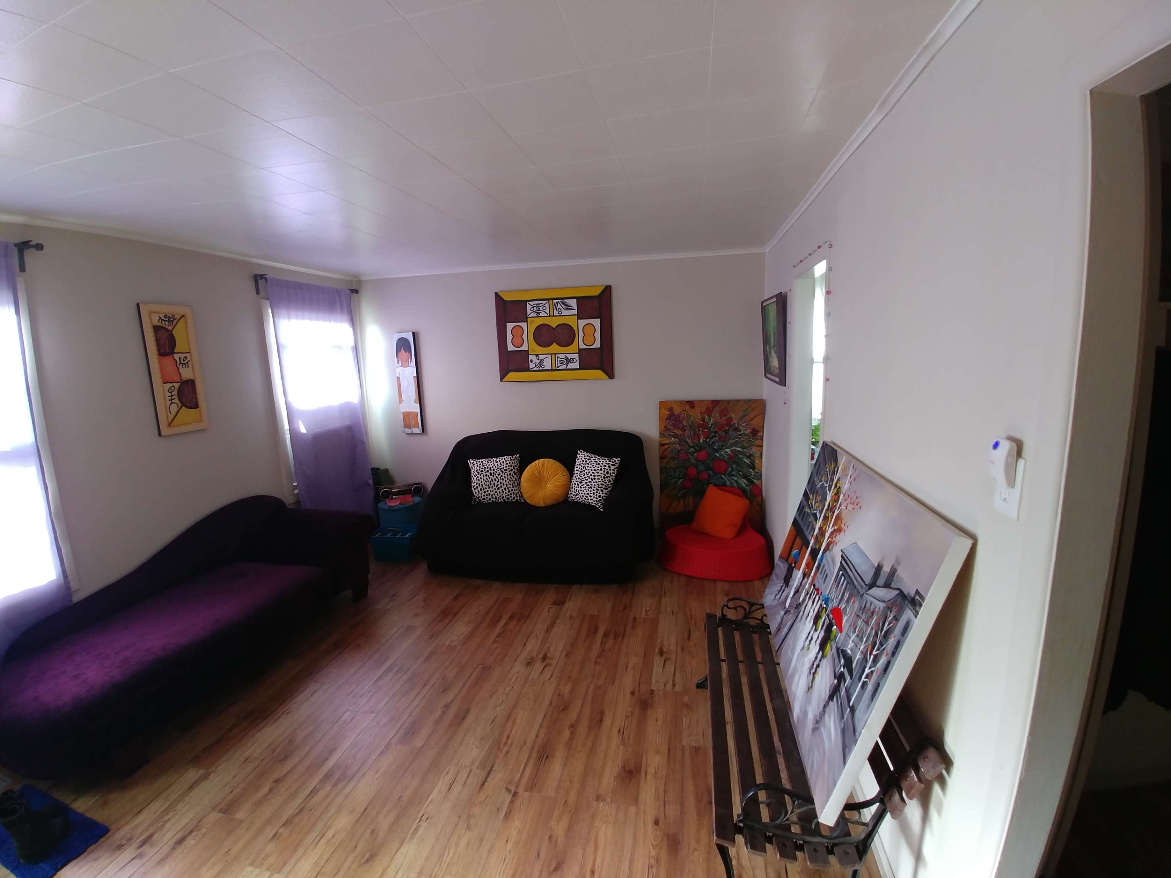 A living room featuring a purple couch, black seating area, colorful artwork on the walls, and a wooden bench near the entrance.