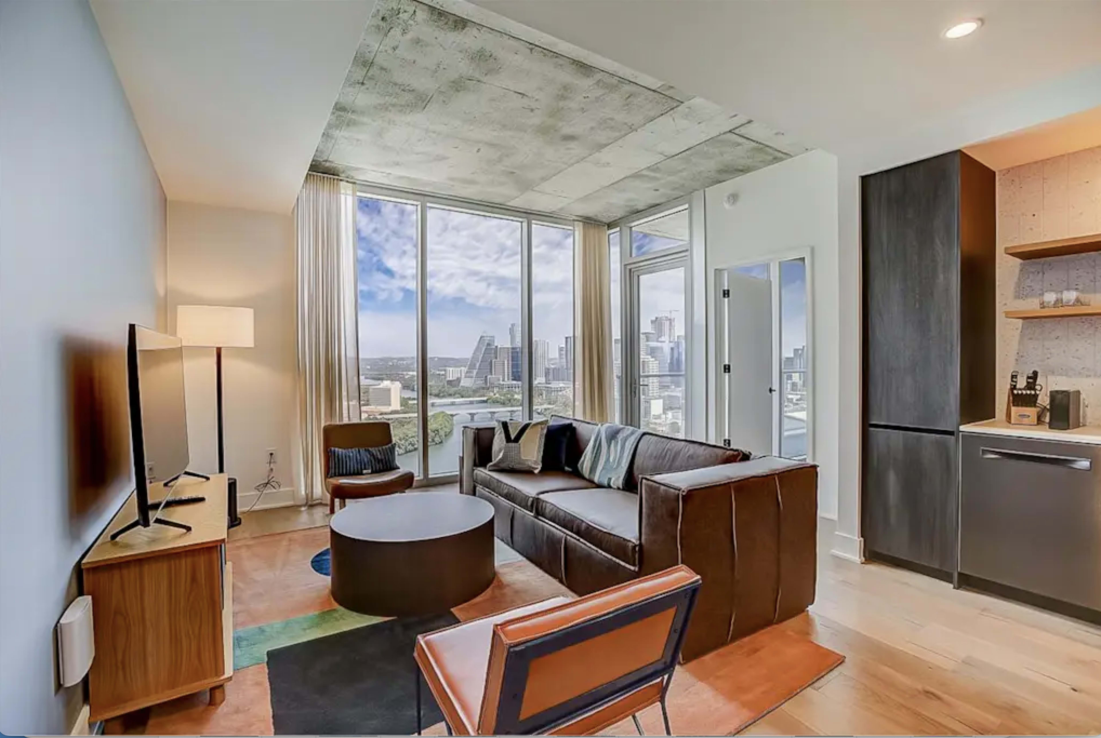 The image shows a modern living room with large windows overlooking a city skyline, featuring a sofa, coffee table, and a TV setup.