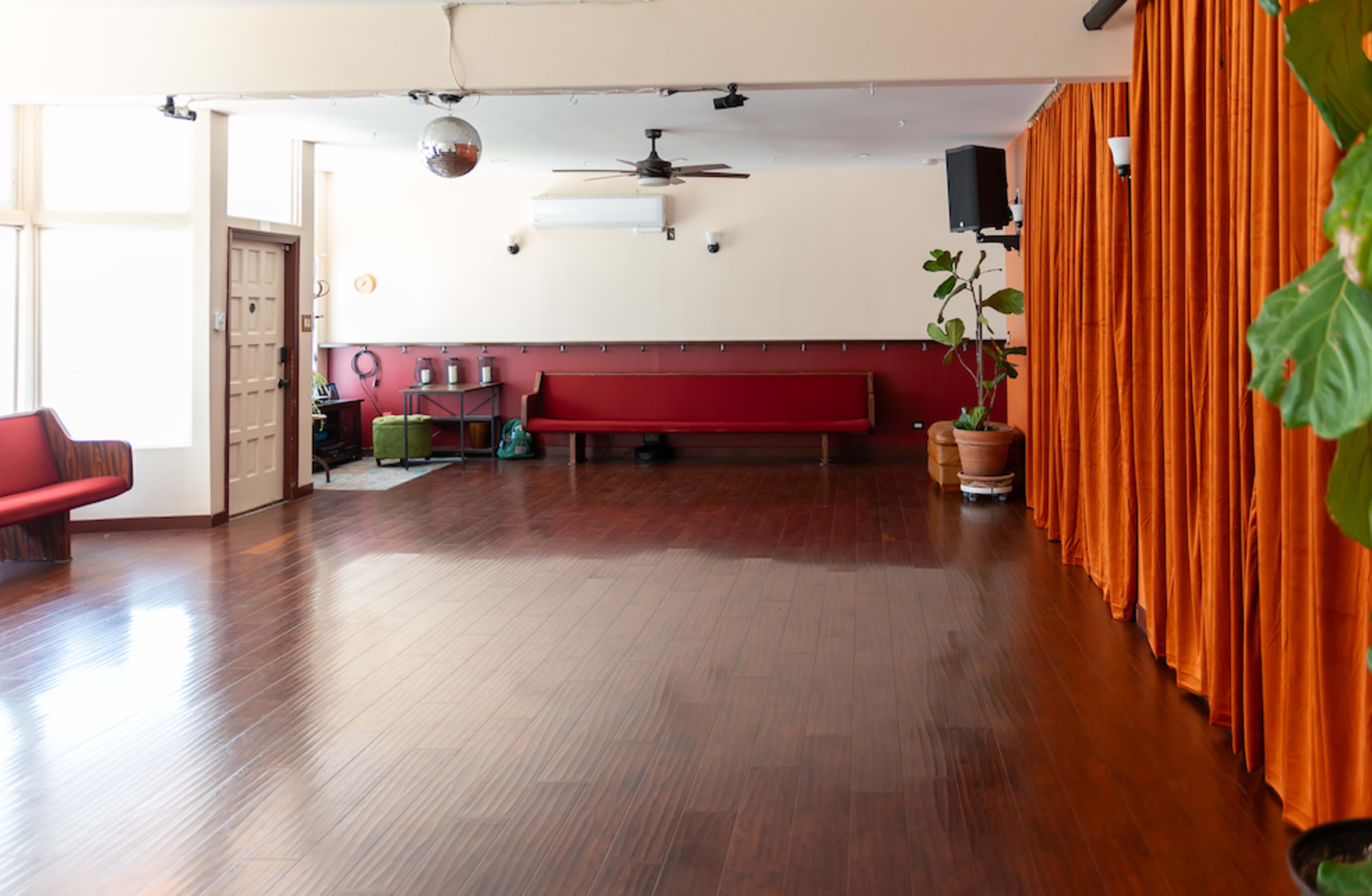 The image shows an empty room with hardwood flooring, a curtain along one wall, and a few plants, along with a red bench and a disco ball hanging from the ceiling.