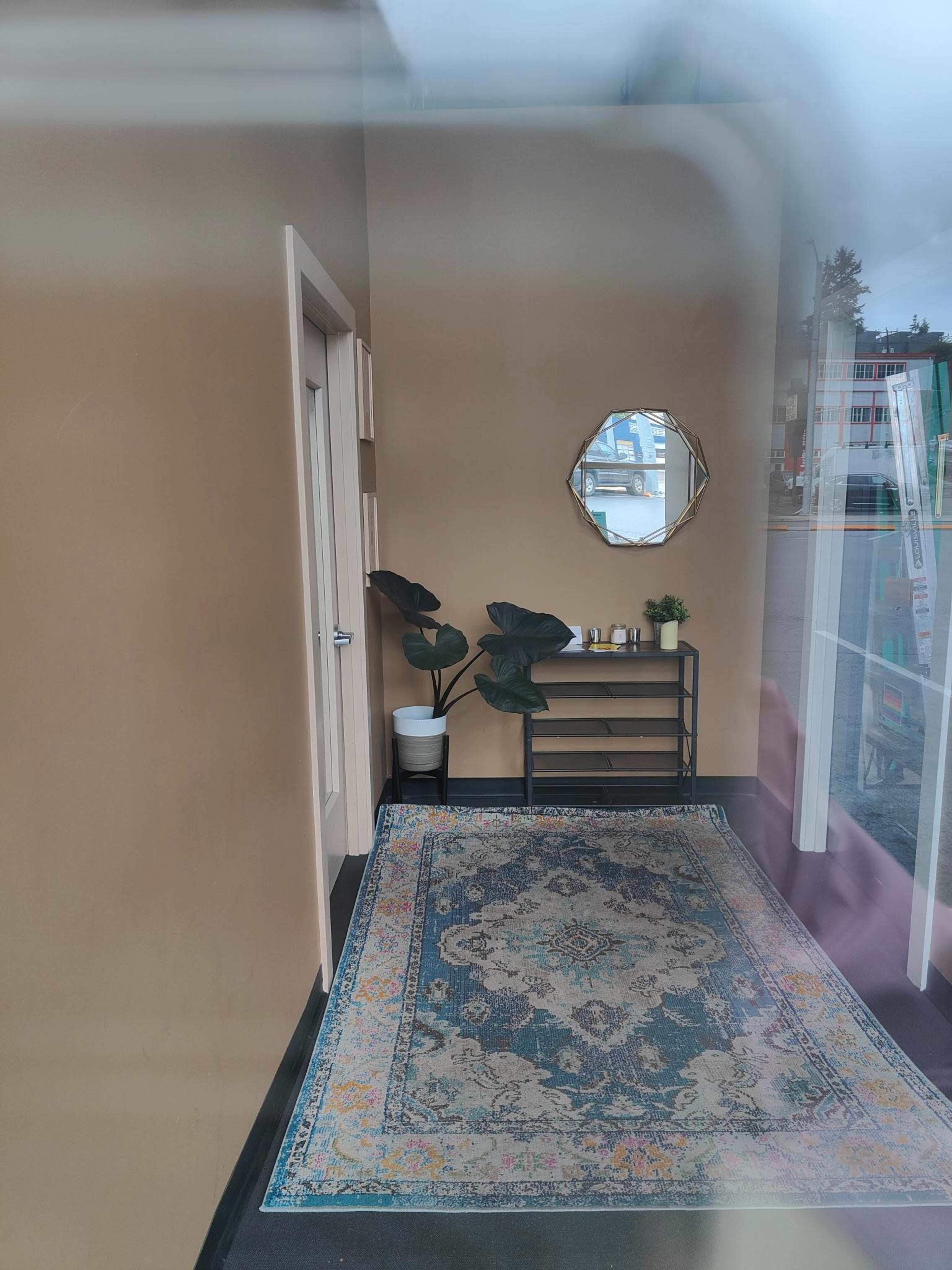 The image shows a small, well-lit hallway featuring a decorative rug, a plant in a pot, a console table with candles, and a round mirror on the wall.