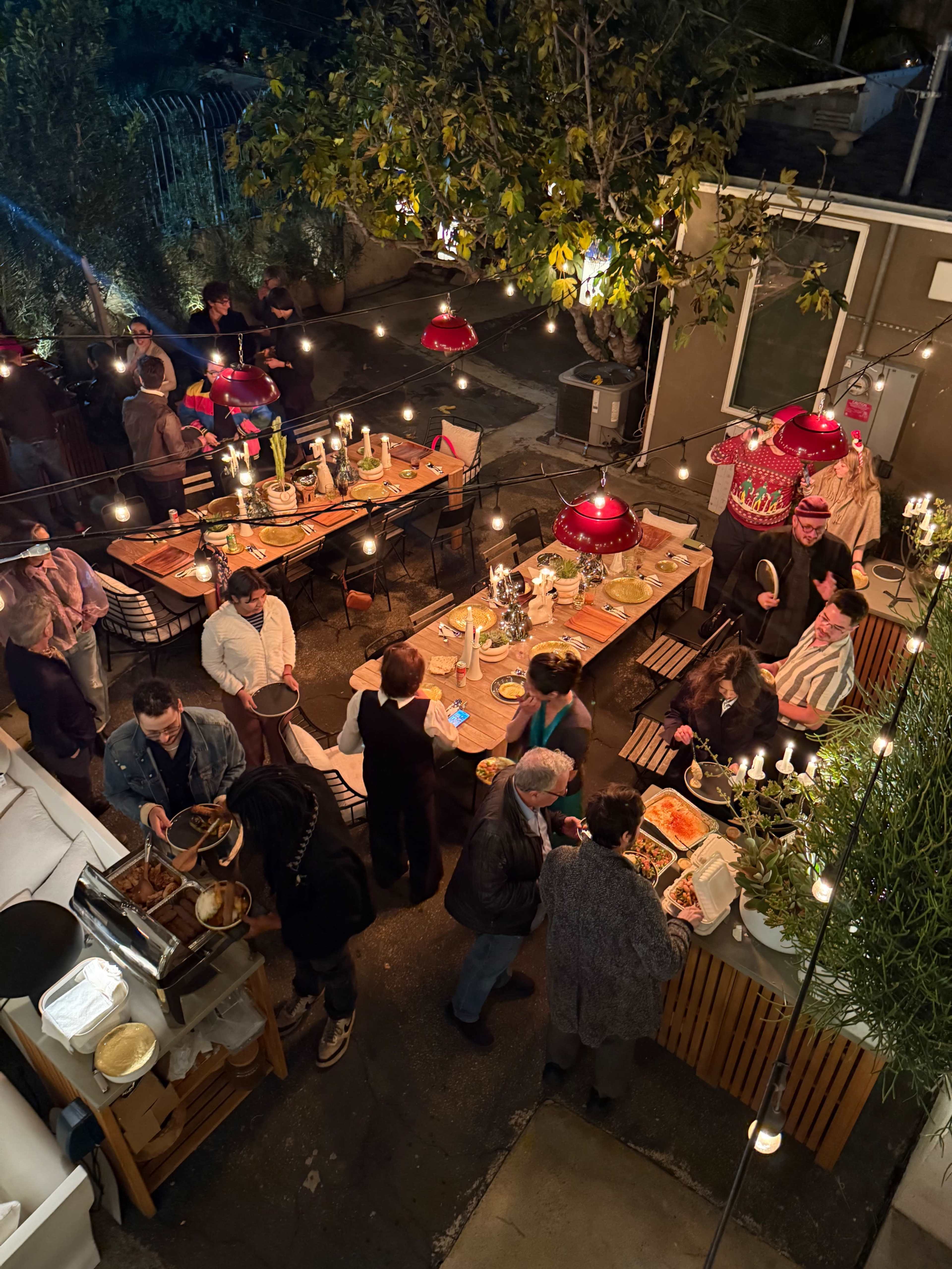 A lively outdoor gathering features guests enjoying a communal dining setup with multiple tables and warmly lit string lights overhead.