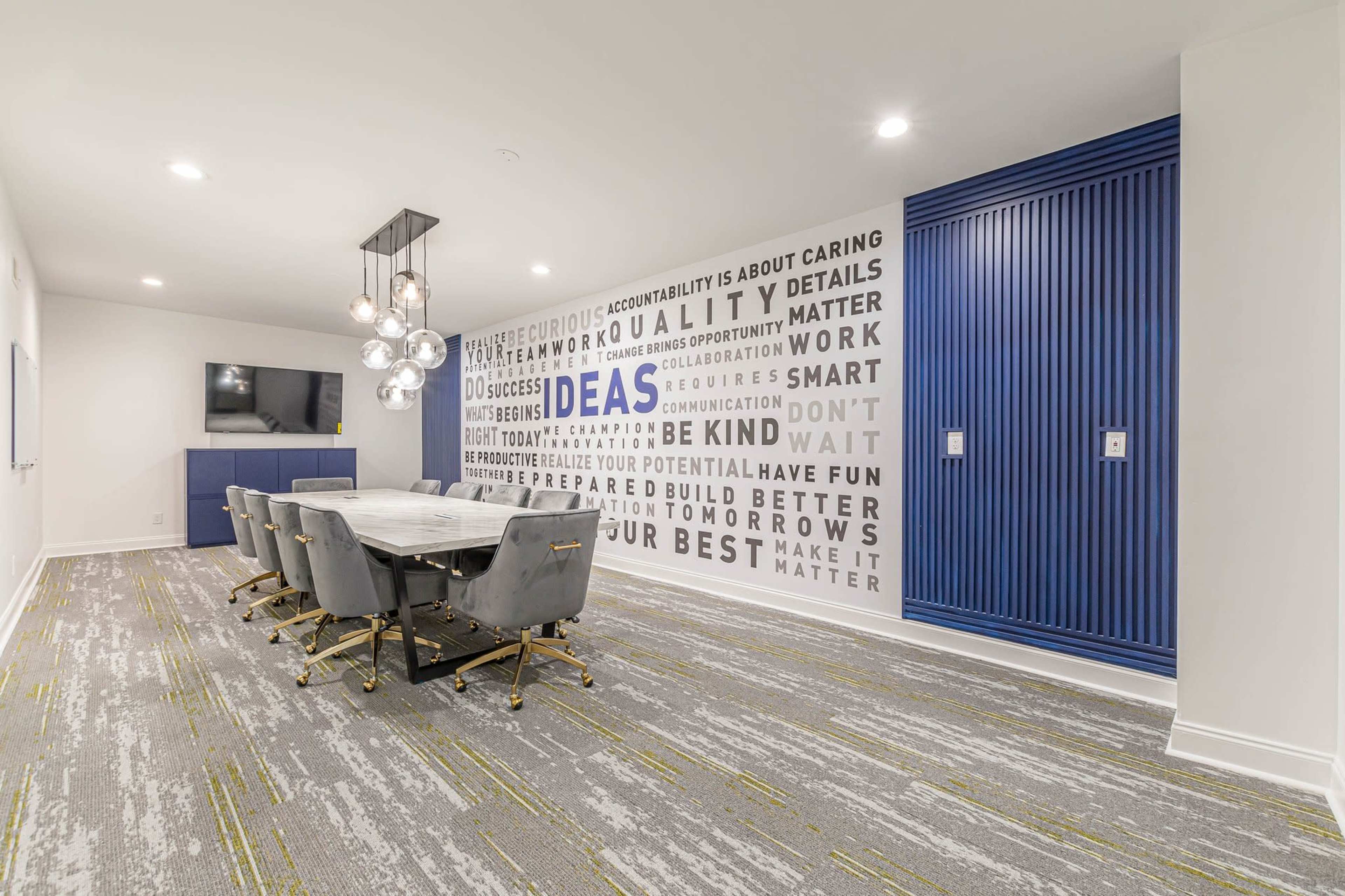 A conference room features a long table surrounded by chairs, with a wall displaying motivational text and a television mounted nearby.