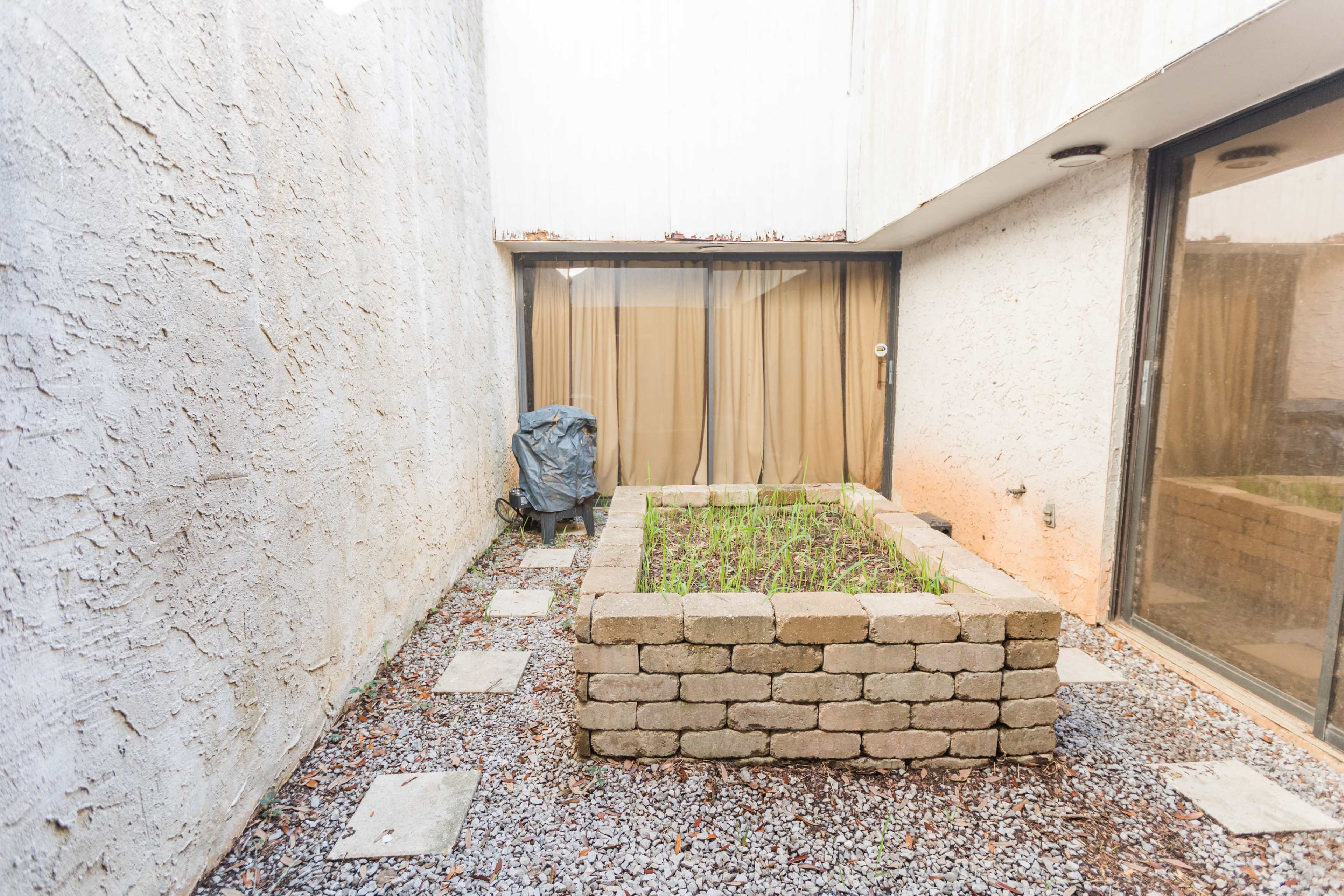 A small courtyard with a raised stone planter bed filled with greenery, surrounded by gravel and concrete walkways.