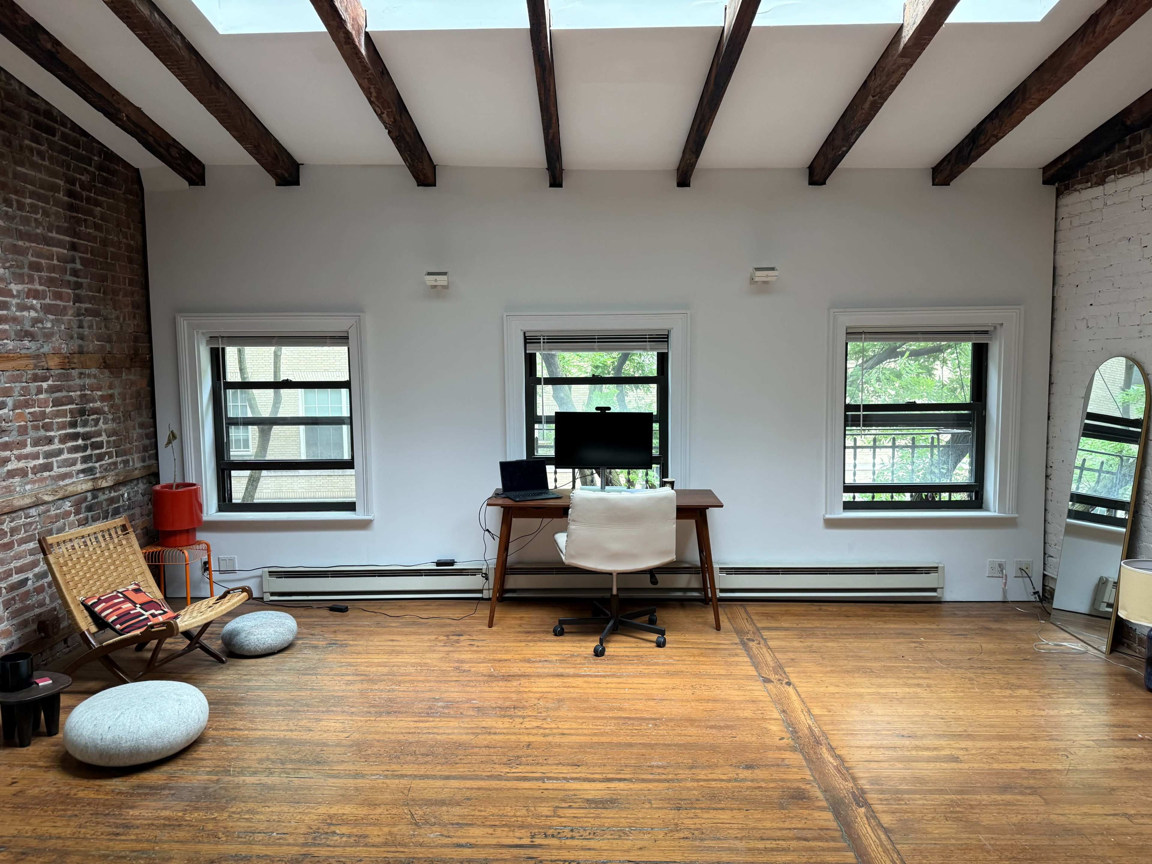 The image shows a spacious room with exposed wooden beams, three windows allowing natural light, a desk with a computer chair, and a seating area featuring a woven chair and floor cushions.