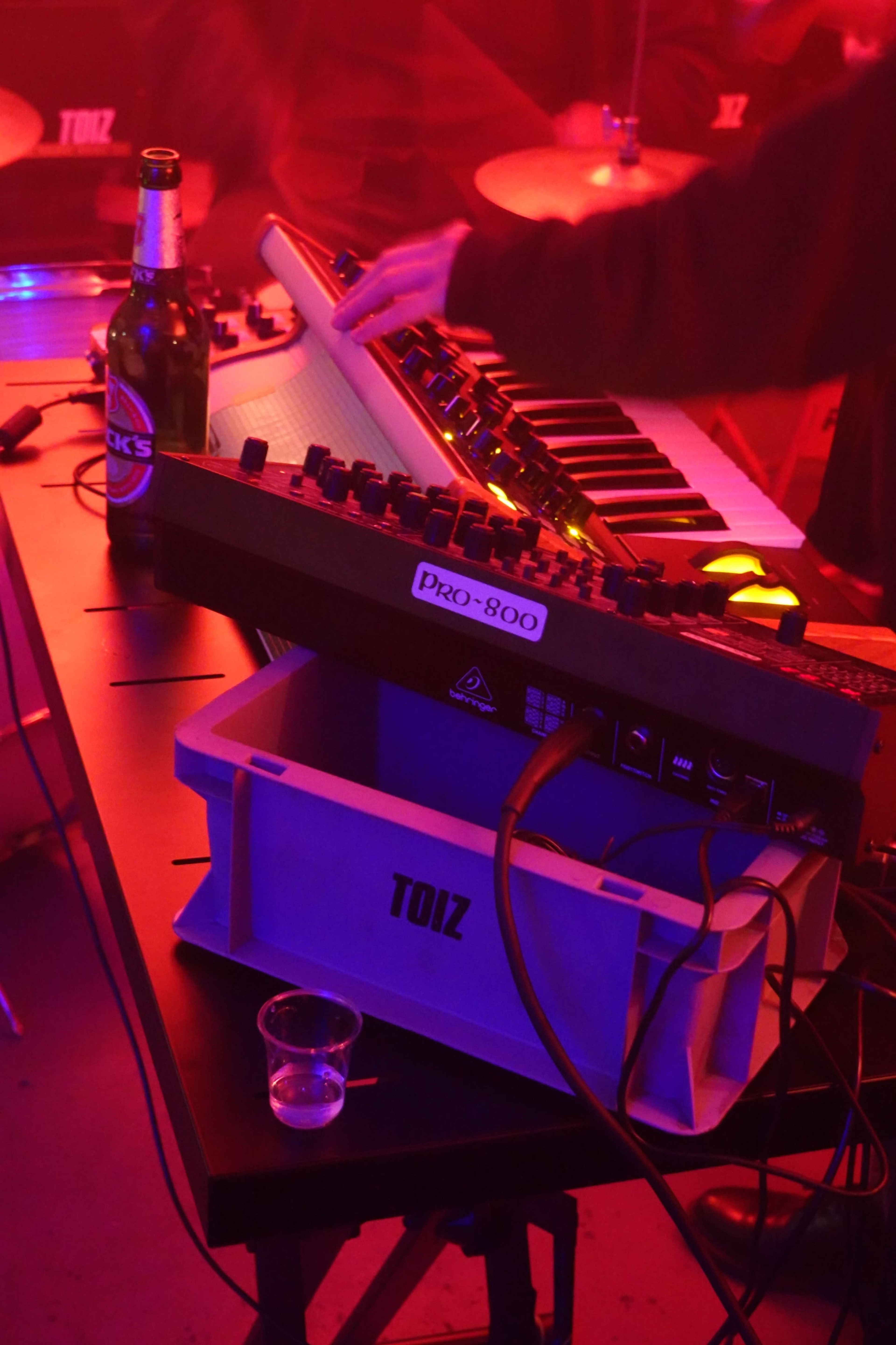 The image shows a close-up of a music setup featuring a synthesizer, a keyboard, a beer bottle, and a small shot glass, all illuminated by red lighting.