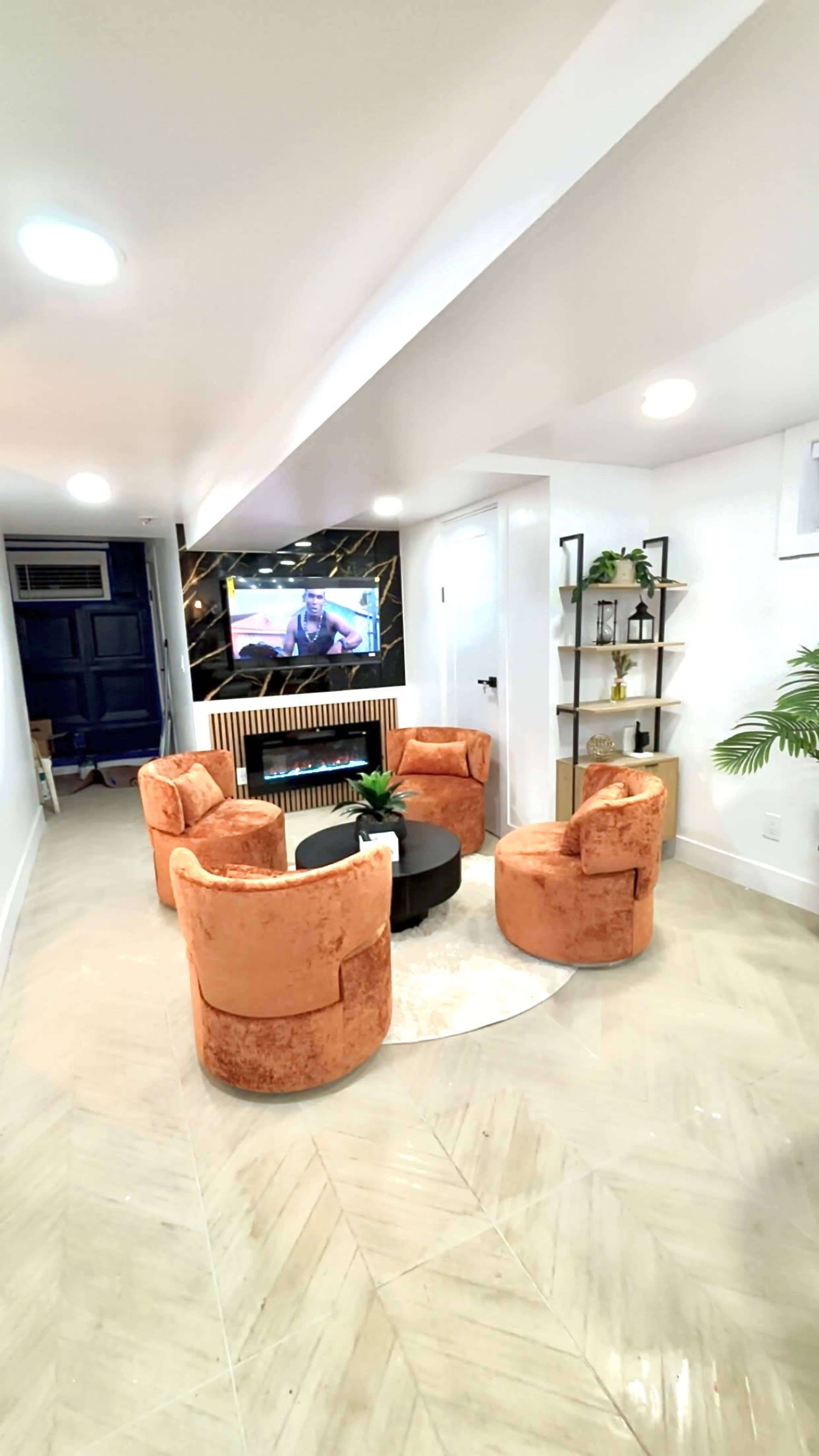 The image shows a modern living room with four orange armchairs arranged around a black coffee table, a television mounted on the wall, and a decorative shelf in the corner.