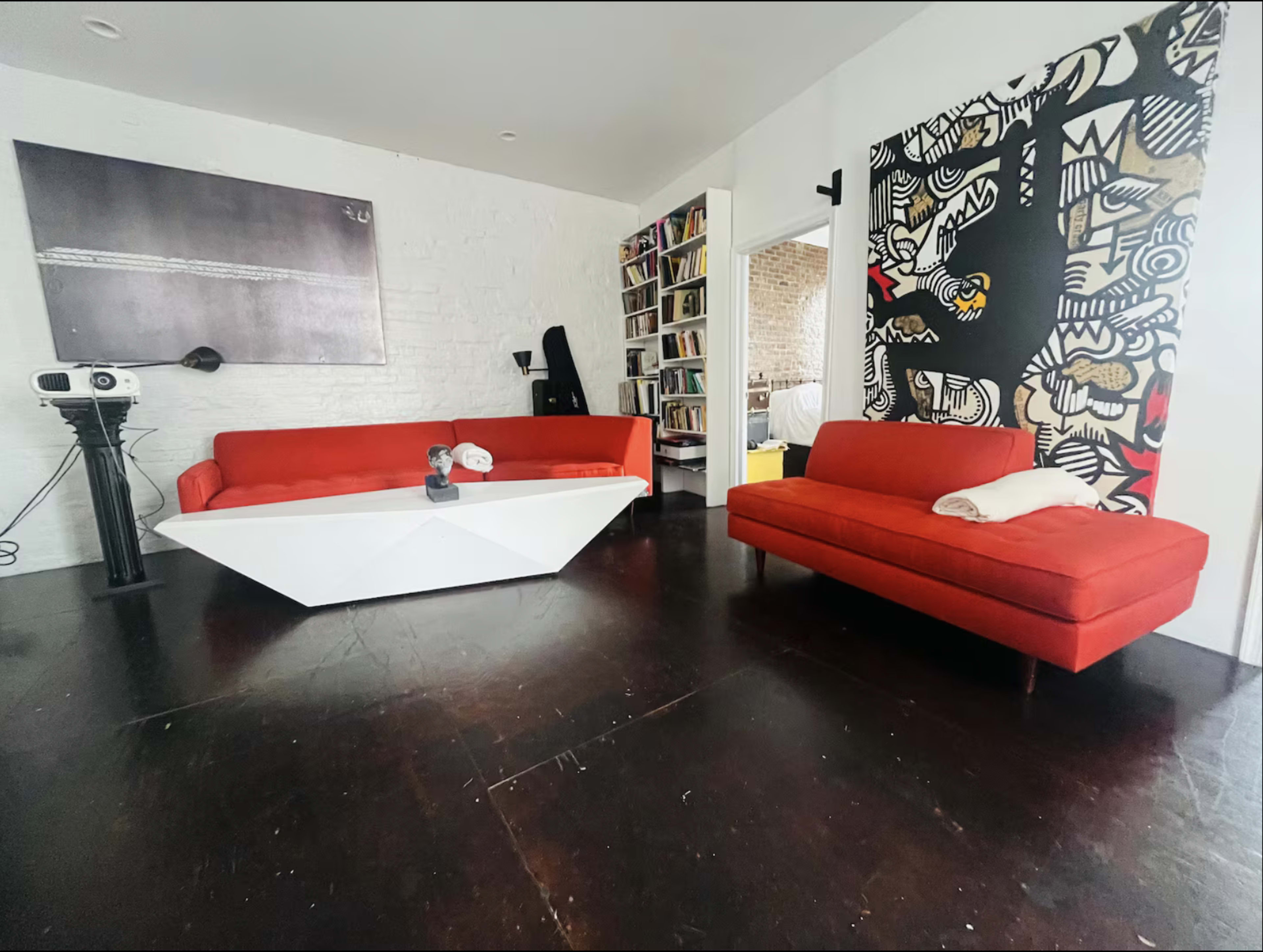 The image shows a modern living room featuring two red sofas, a white angular coffee table, and a large wall art piece on one side.