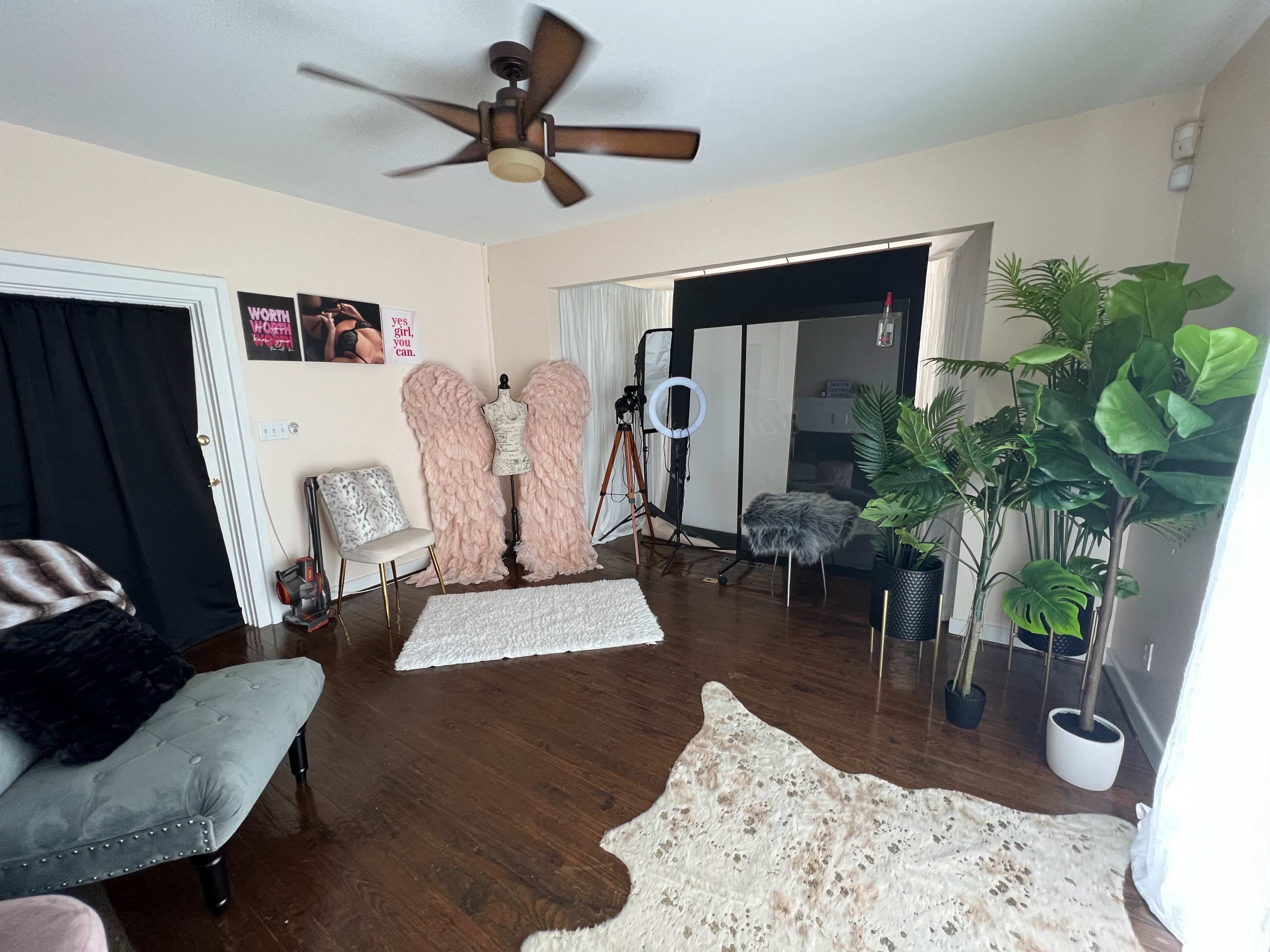 A well-lit room featuring large faux fur angel wings, a clothing rack, a ring light, potted plants, and a soft area rug on a wooden floor.