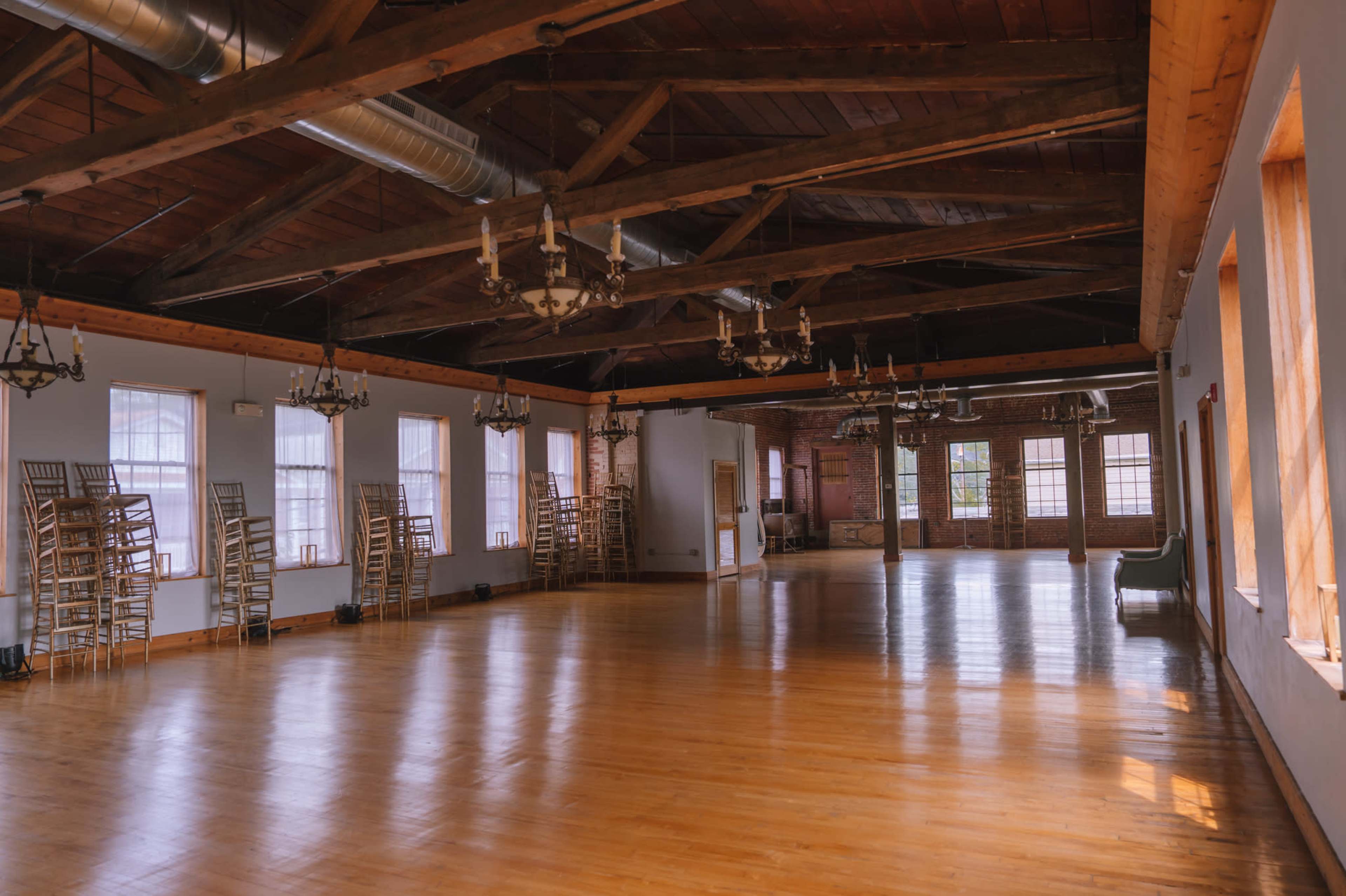 The image shows a spacious, empty room with wooden floors, exposed beams, and several tall windows, featuring stacked chairs against the walls.