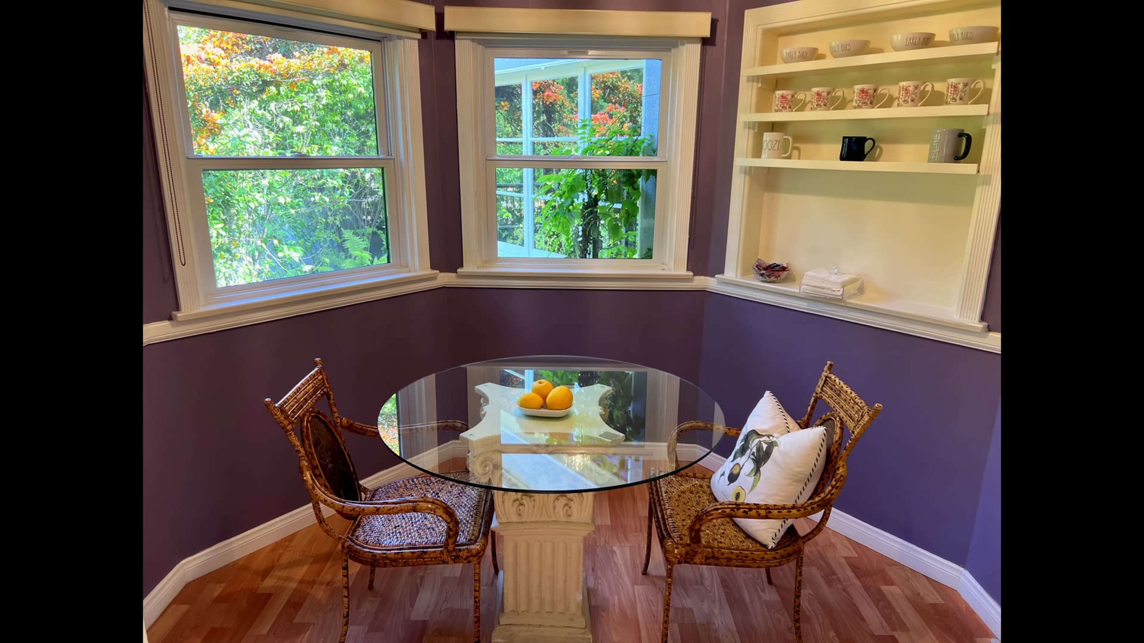 A round glass table with two wicker chairs is set in a corner nook, surrounded by windows and adorned with a centerpiece of oranges.