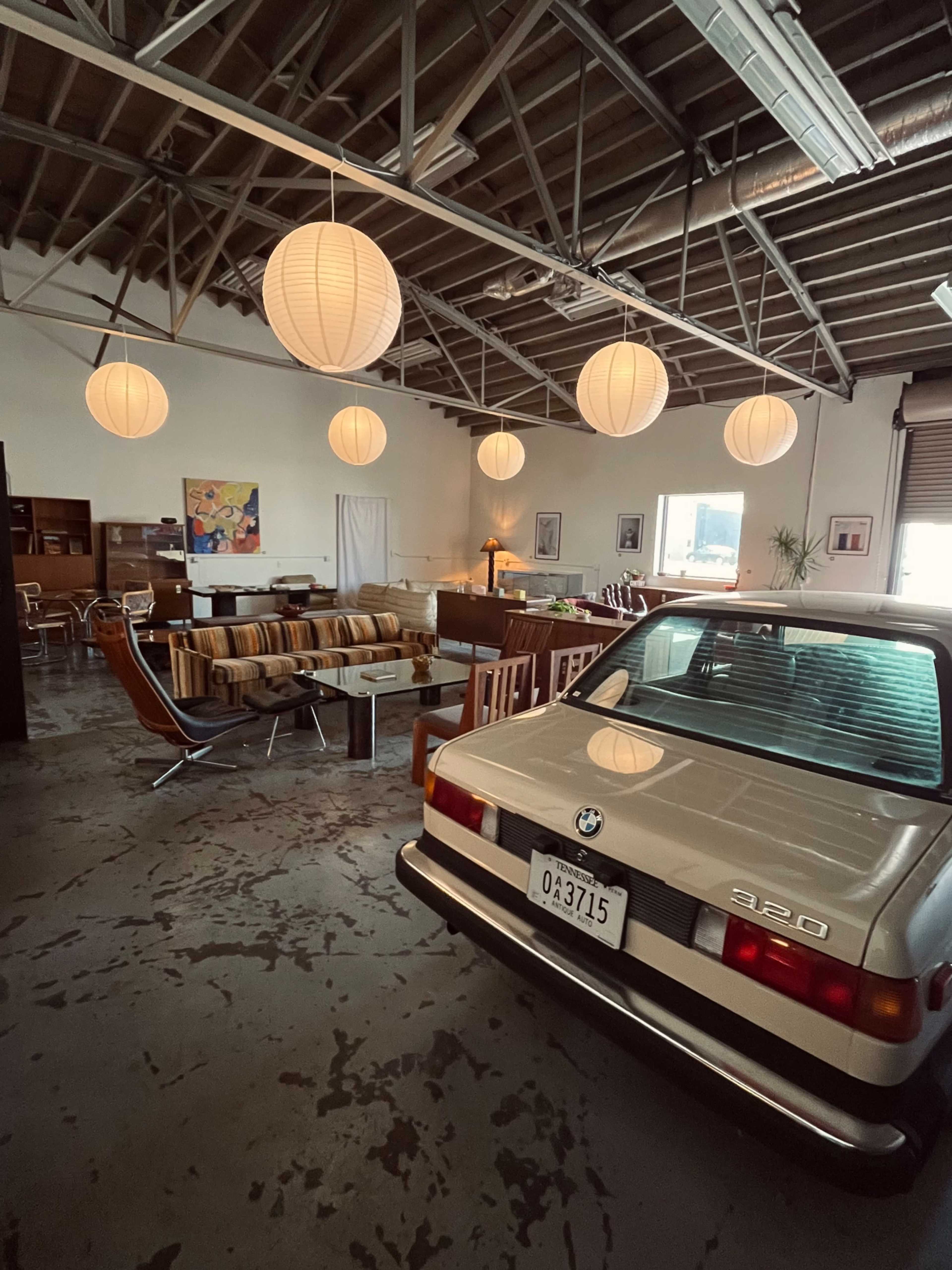 An industrial-style loft features a vintage BMW parked inside, surrounded by modern furniture and suspended paper lanterns.
