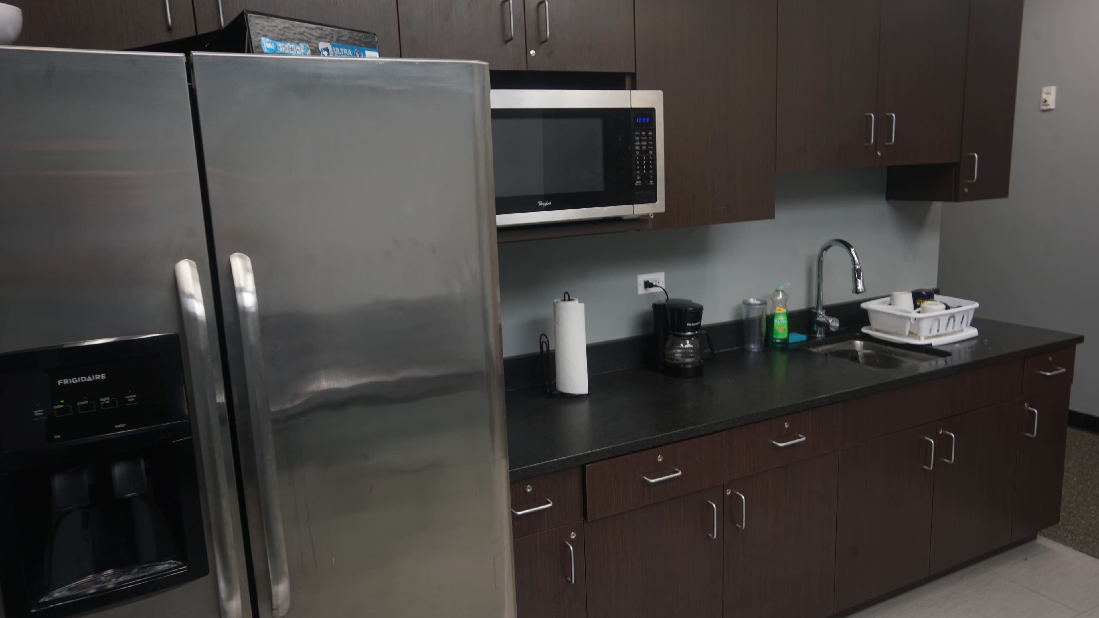 A stainless steel refrigerator and microwave are installed in a kitchen with dark wood cabinets and a black countertop.