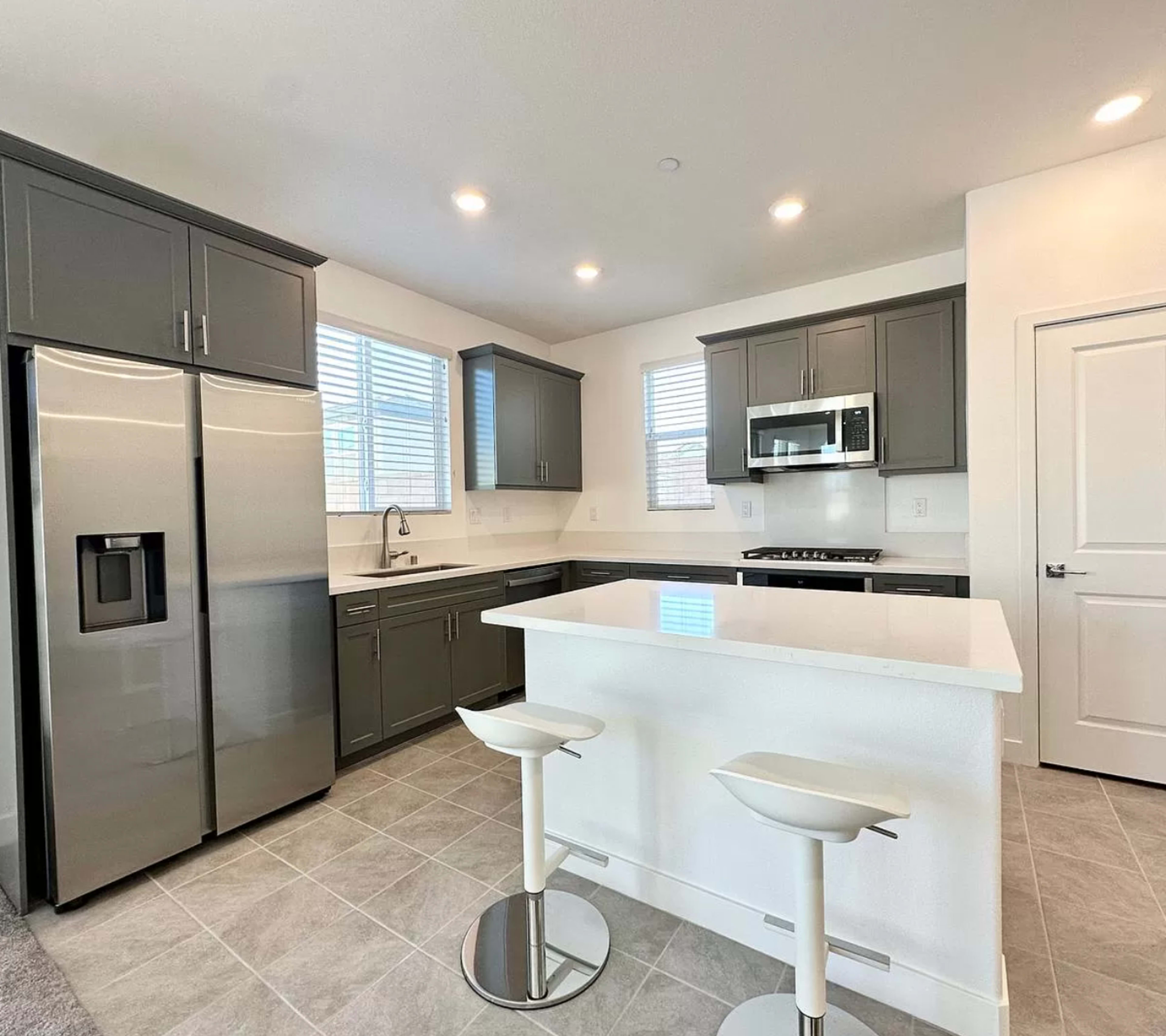 A modern kitchen features gray cabinetry, stainless steel appliances, and a white island with two bar stools.