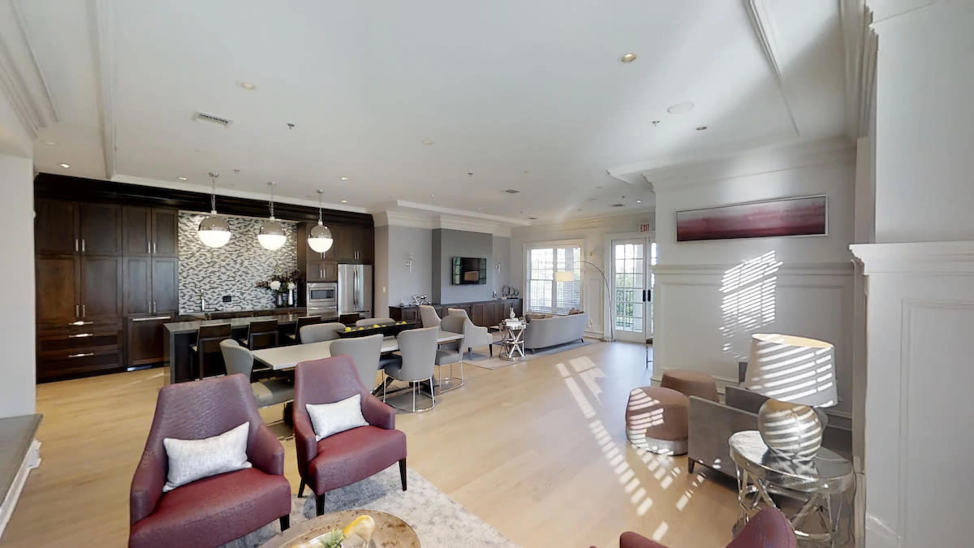 The image shows a modern living area with a kitchen, featuring seating arrangements and large windows that allow natural light to enter.