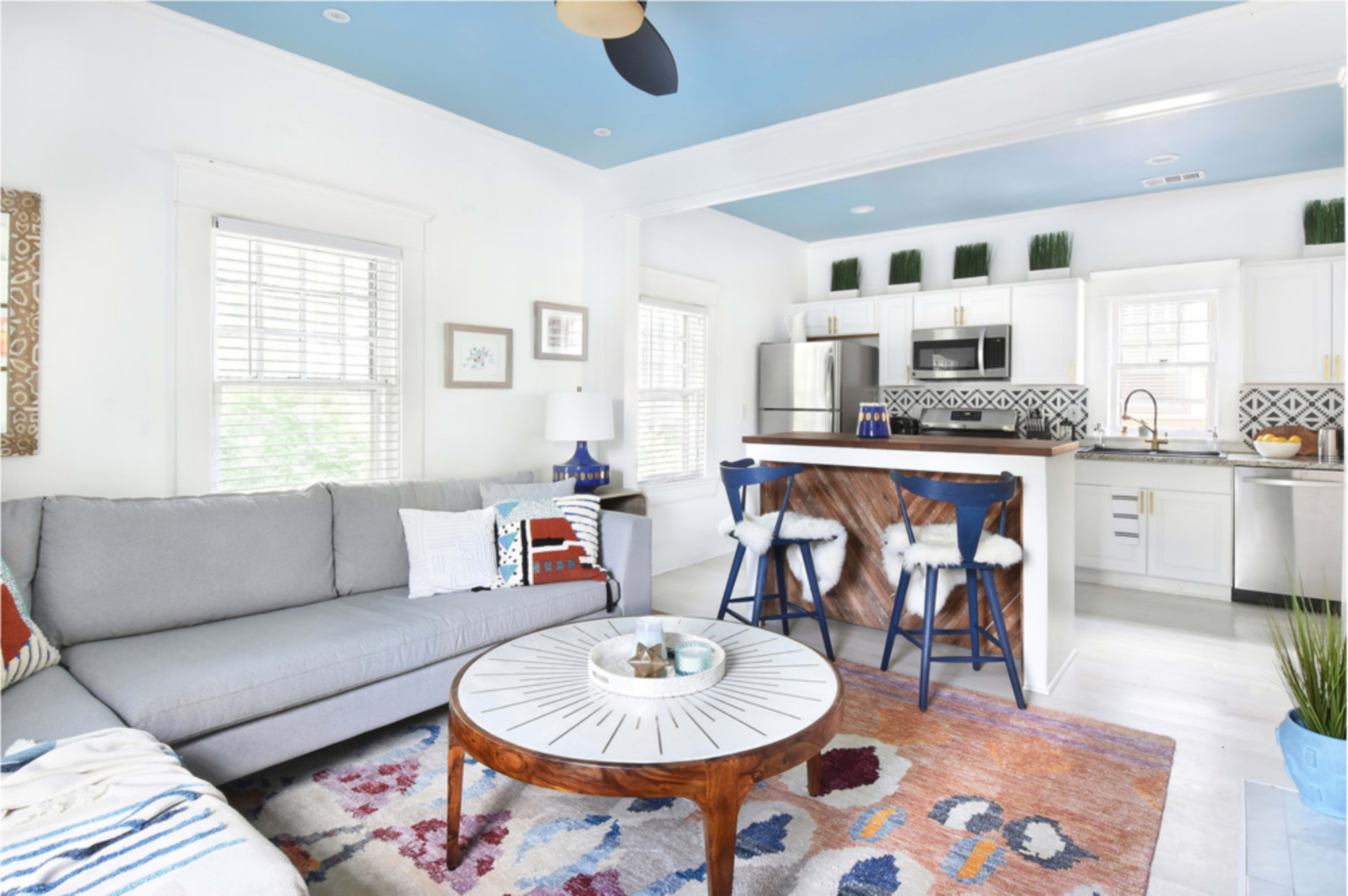 A modern living room features a gray sofa, a round coffee table with a sunburst design, and a kitchen area with white cabinets and patterned tile.