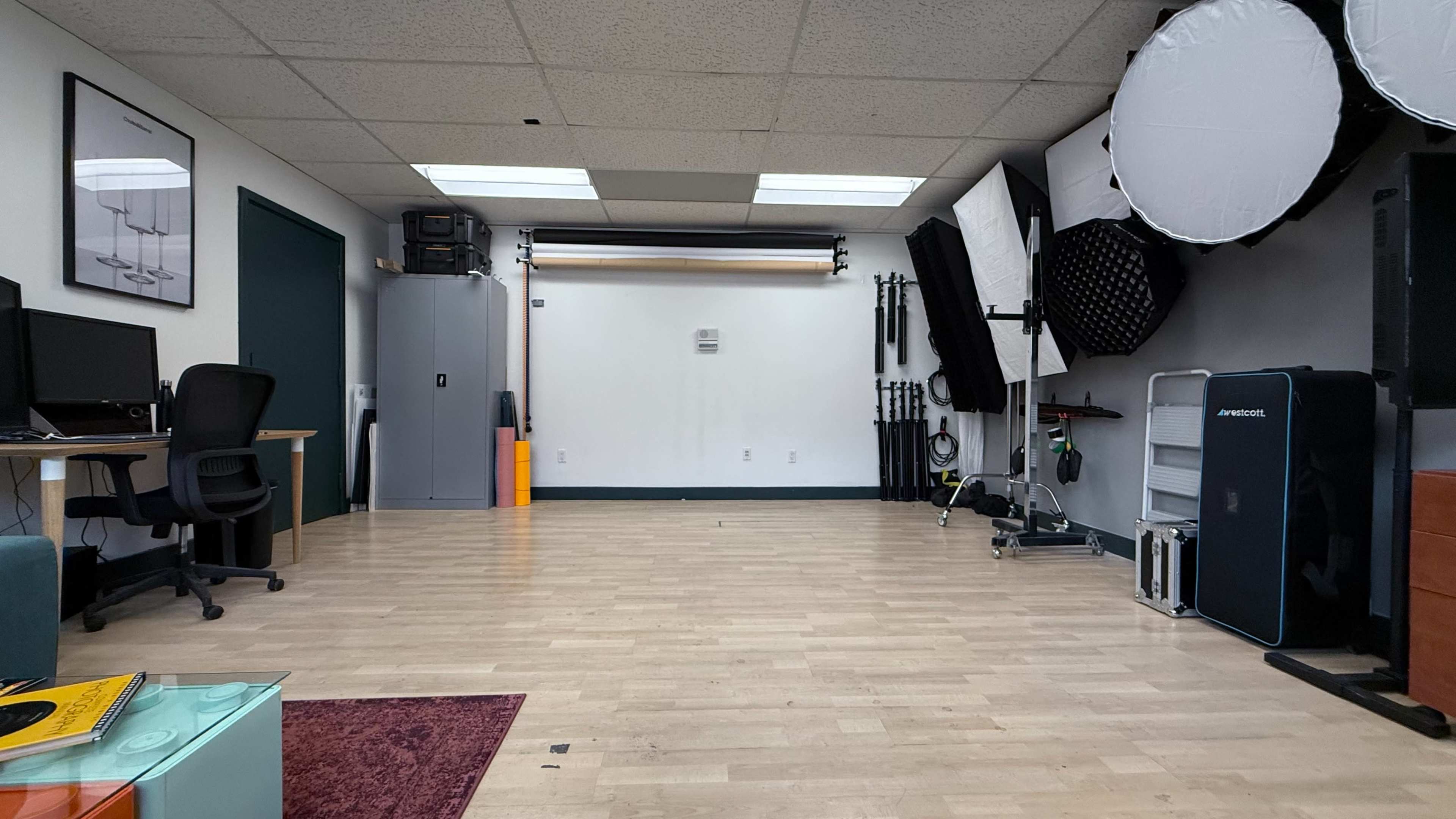 The image shows a spacious studio with a wooden floor, two computer workstations, and various lighting equipment arranged along the walls.