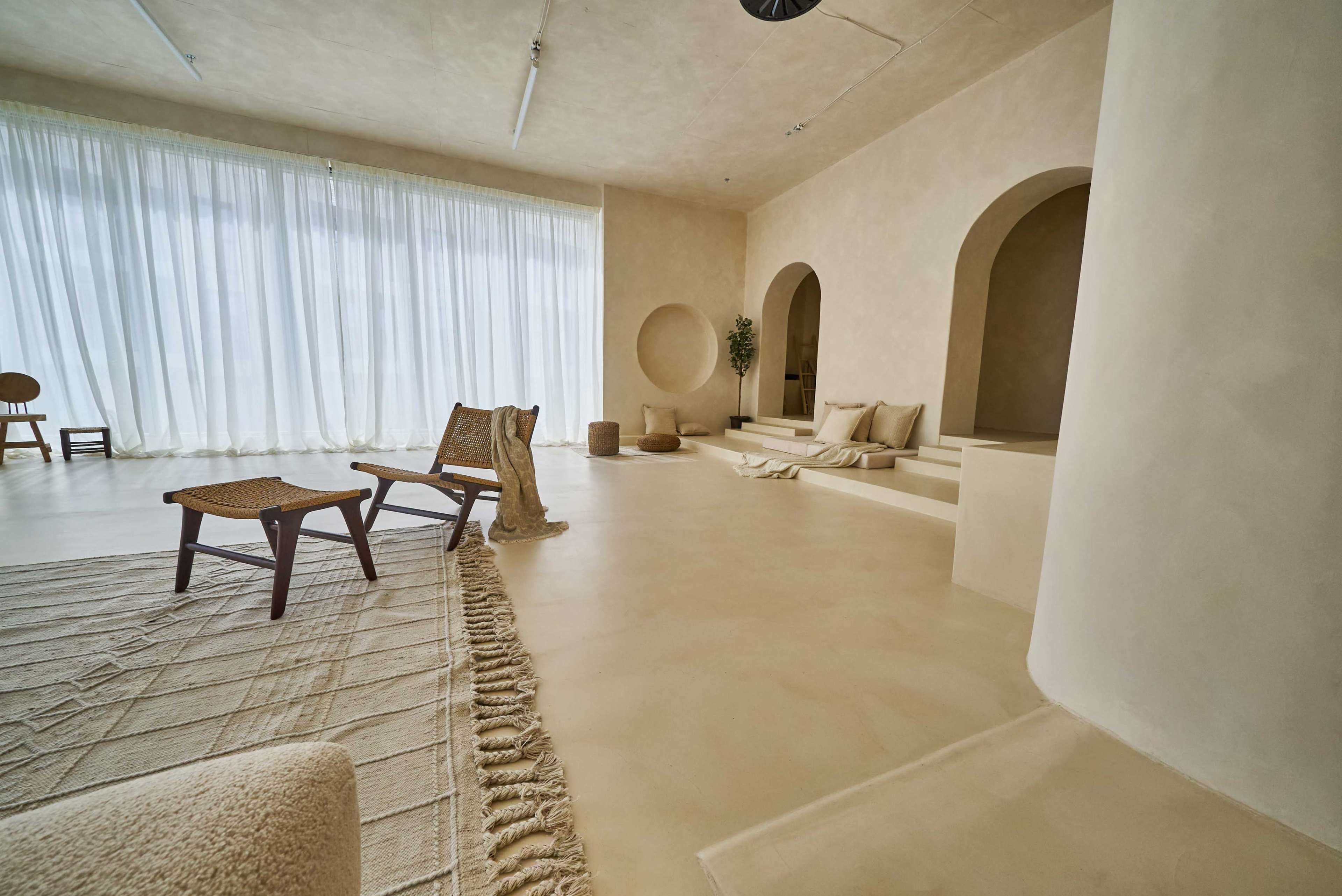 The image shows a minimalistic interior space with light-colored walls, large windows covered by sheer curtains, and various seating arrangements including a woven chair and a few cushions on the floor.