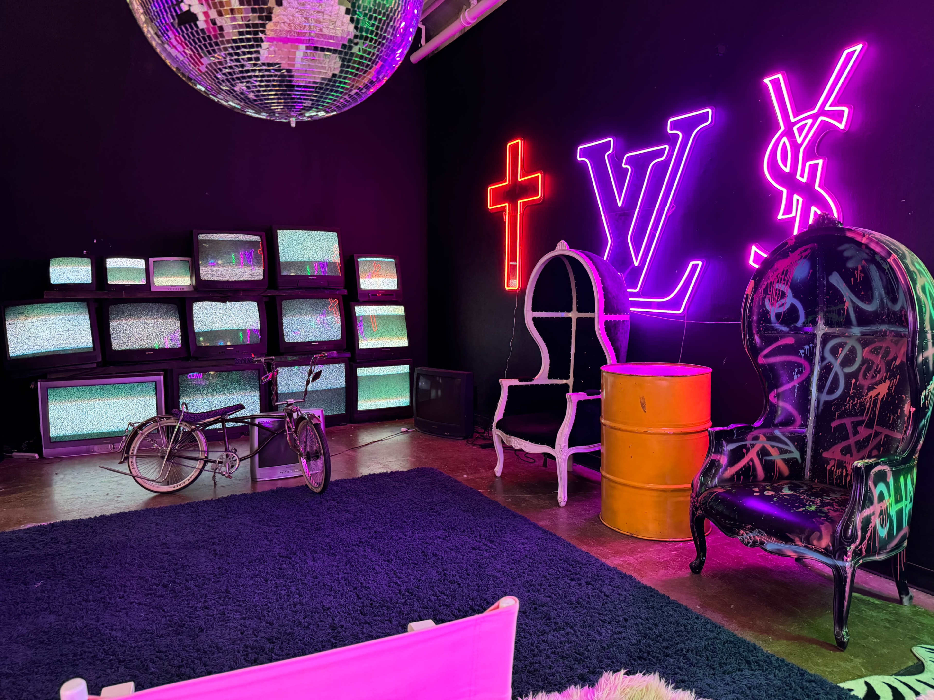 The scene features a dimly lit room with a disco ball, neon logos on the wall, several vintage television sets displaying static, graffiti-covered furniture, and a bicycle.