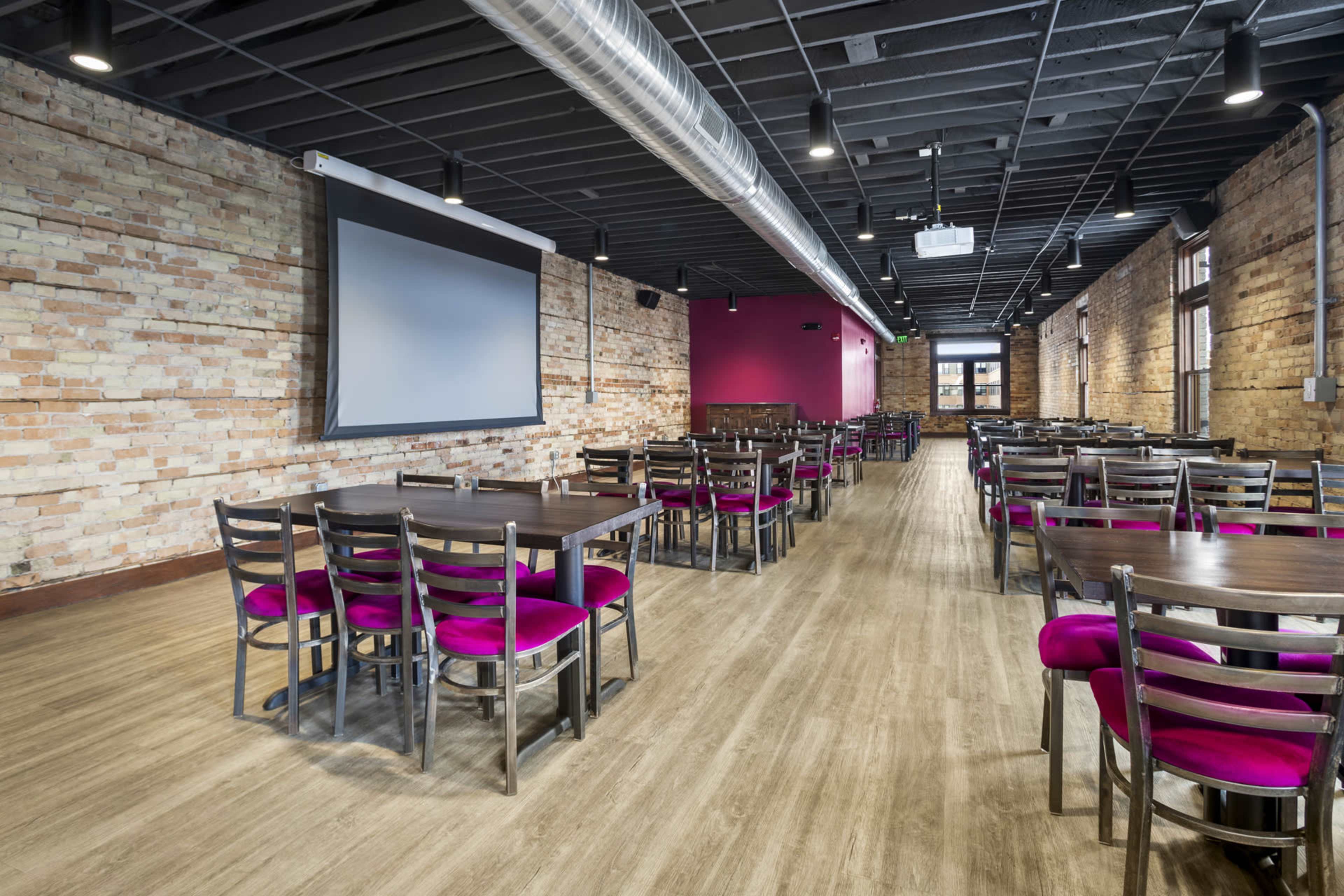 The image shows a spacious restaurant interior with rows of tables and chairs, featuring exposed brick walls and a large screen on one wall.
