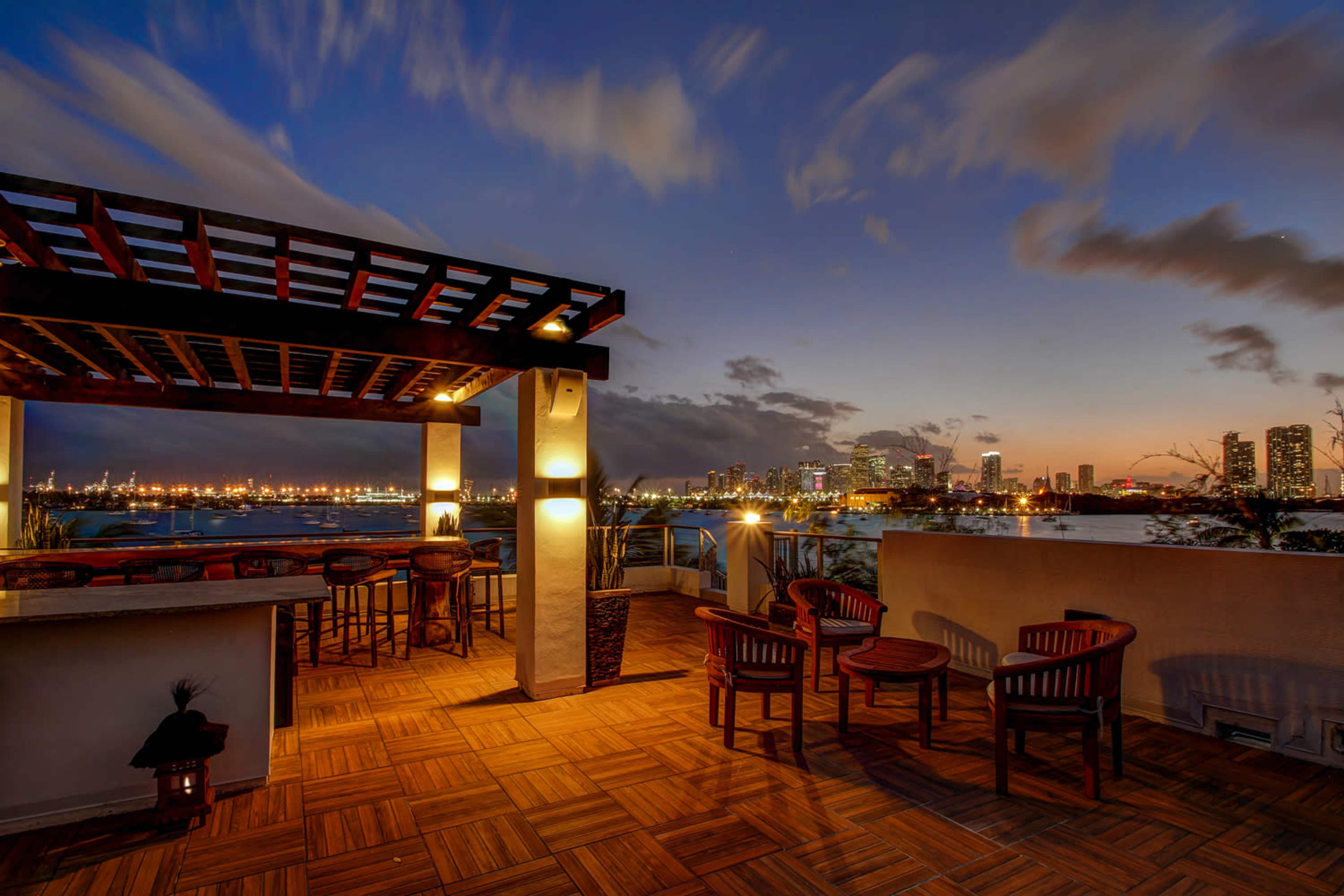 The image shows a rooftop terrace with wooden furniture and a view of a city skyline during sunset.