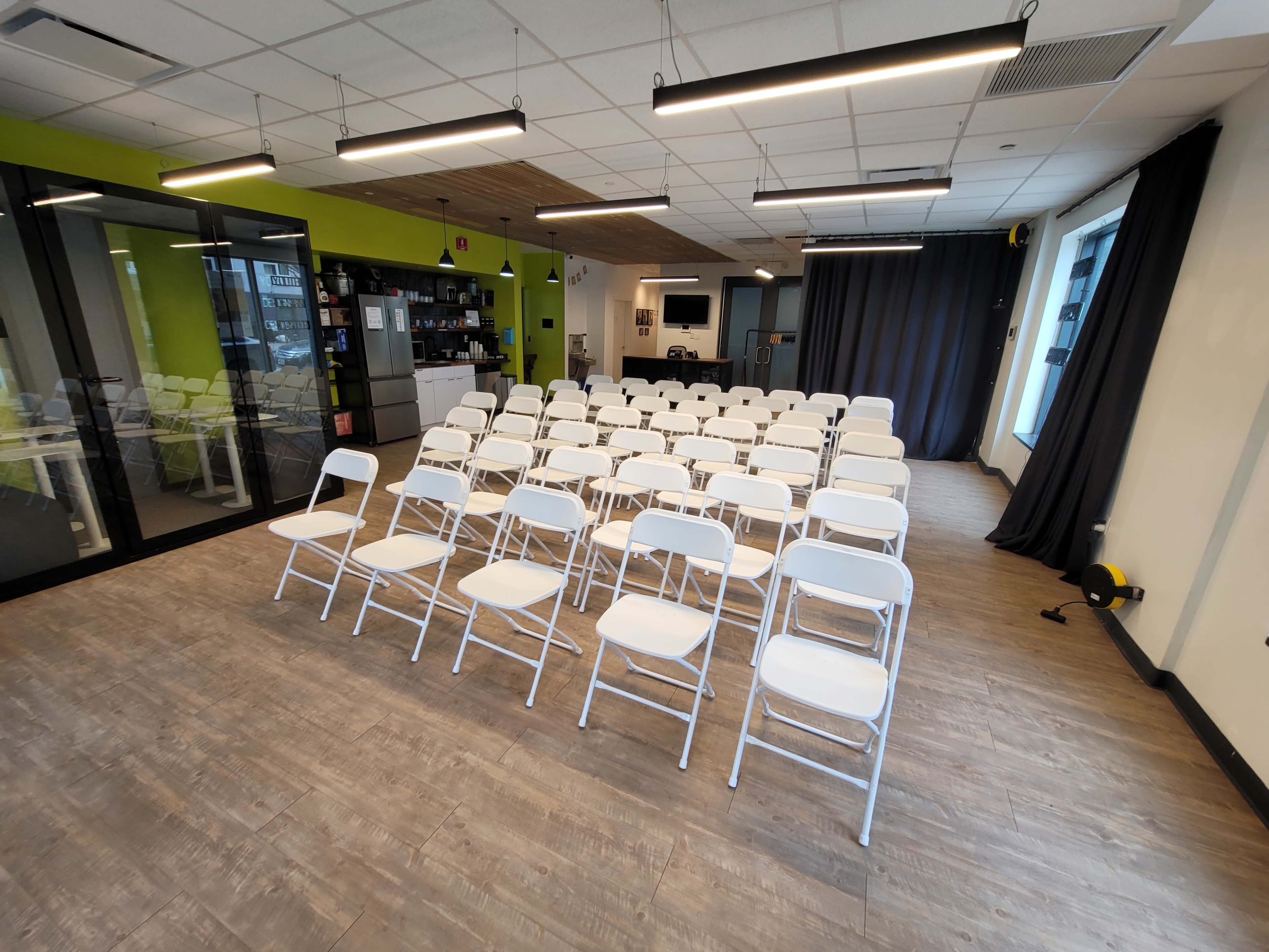The image shows a row of white folding chairs arranged in front of a wall of windows in a modern room with light wood flooring.