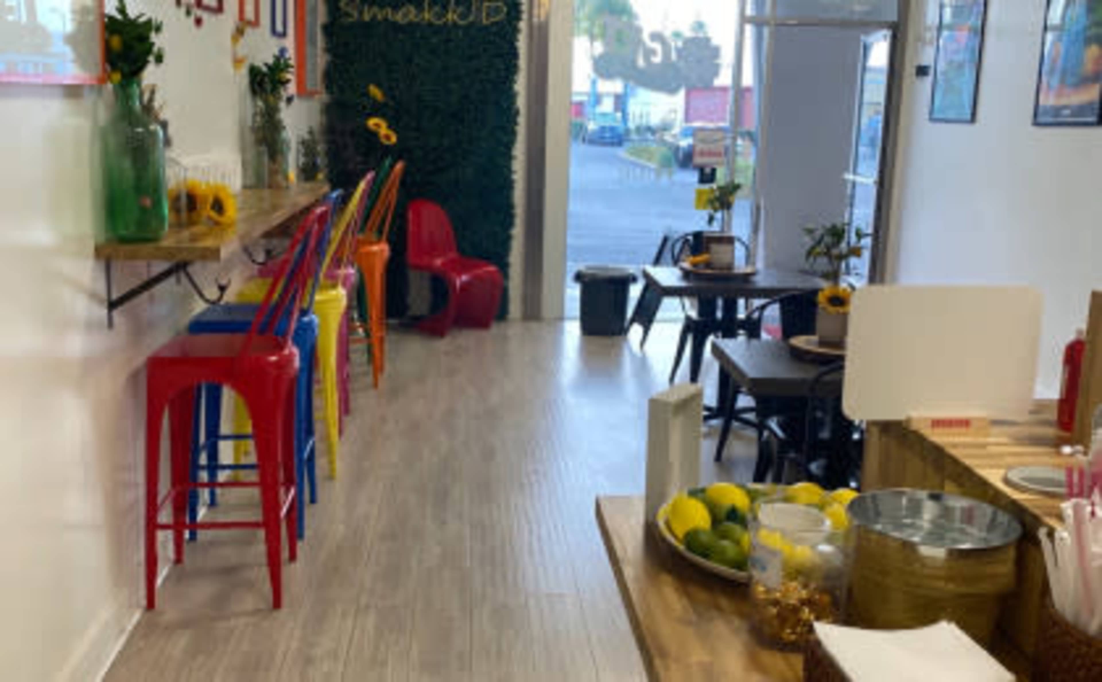 The image shows a brightly decorated café interior with colorful chairs, wooden tables, and a counter displaying various items, including fruit and utensils.