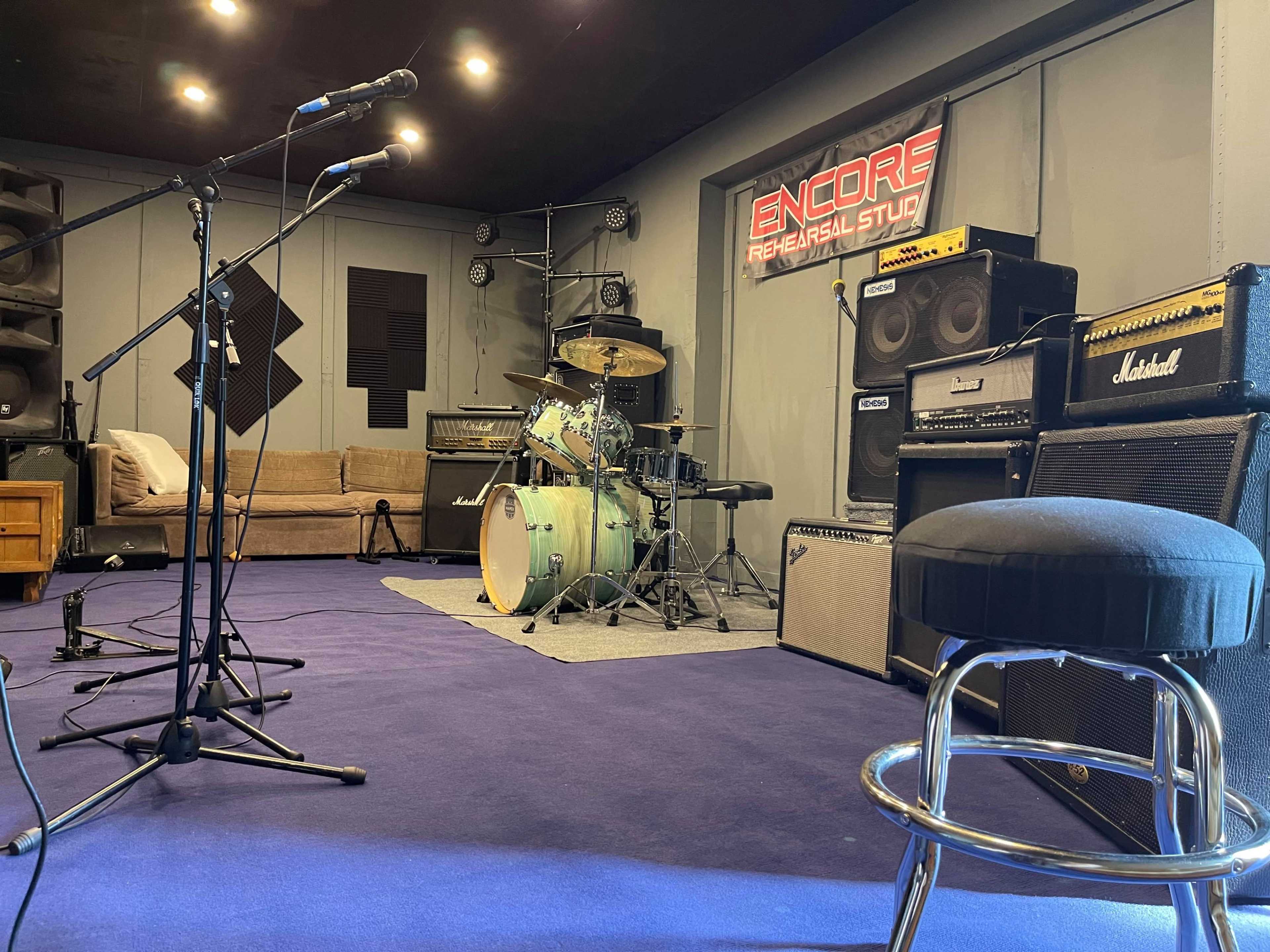 A rehearsal studio features microphones, a drum set, amplifiers, and a seating area.