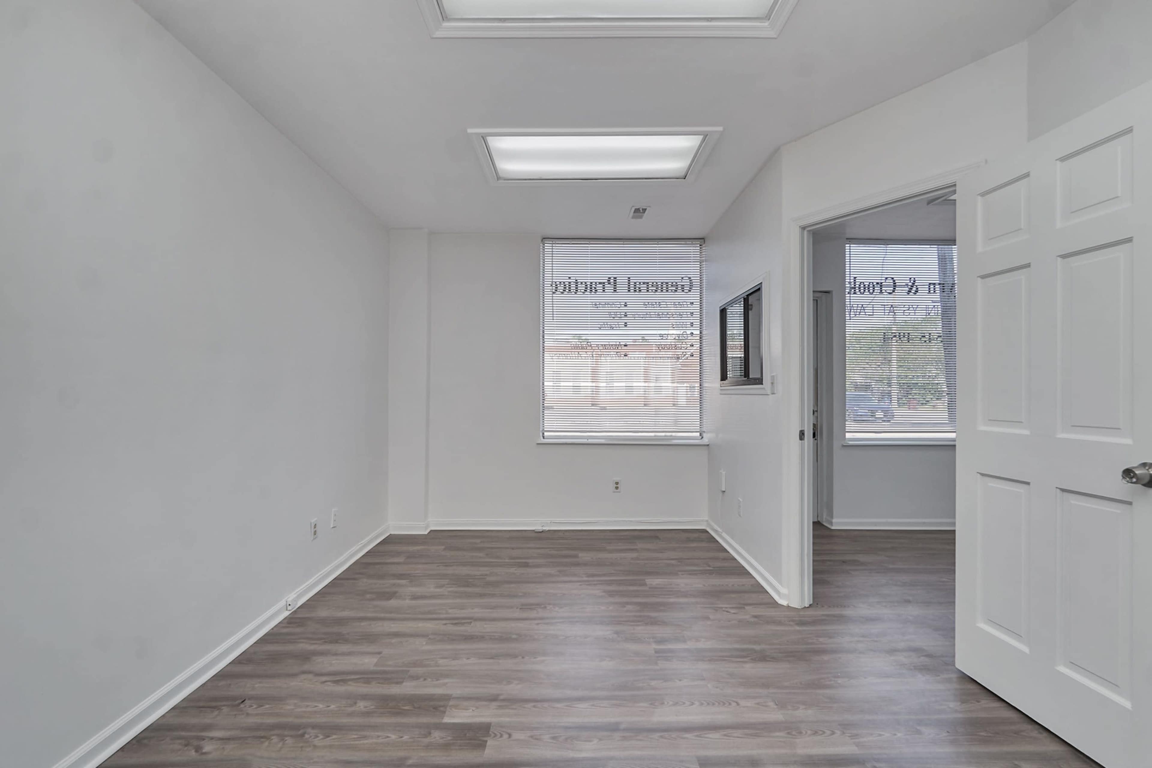 A vacant office space with white walls, hardwood-like flooring, and a door leading to another room.