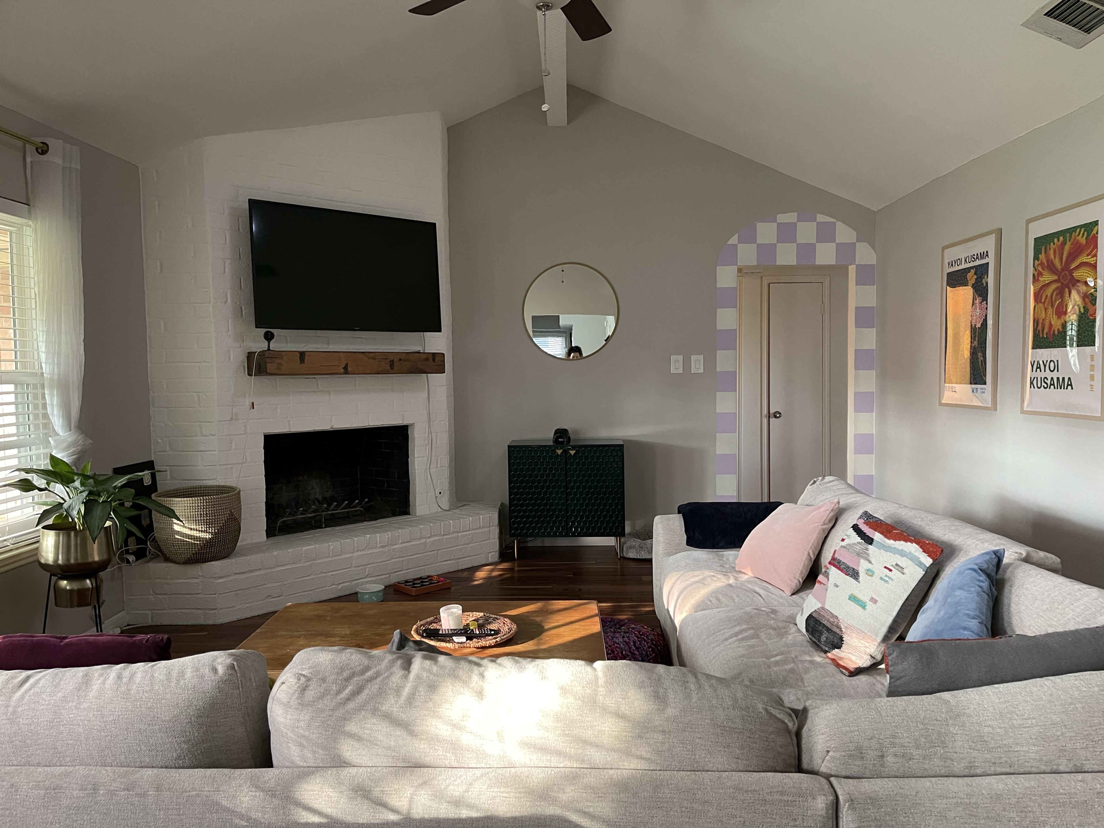 A living room featuring a gray sectional sofa, a white brick fireplace with a mounted TV, and colorful artwork on the wall.