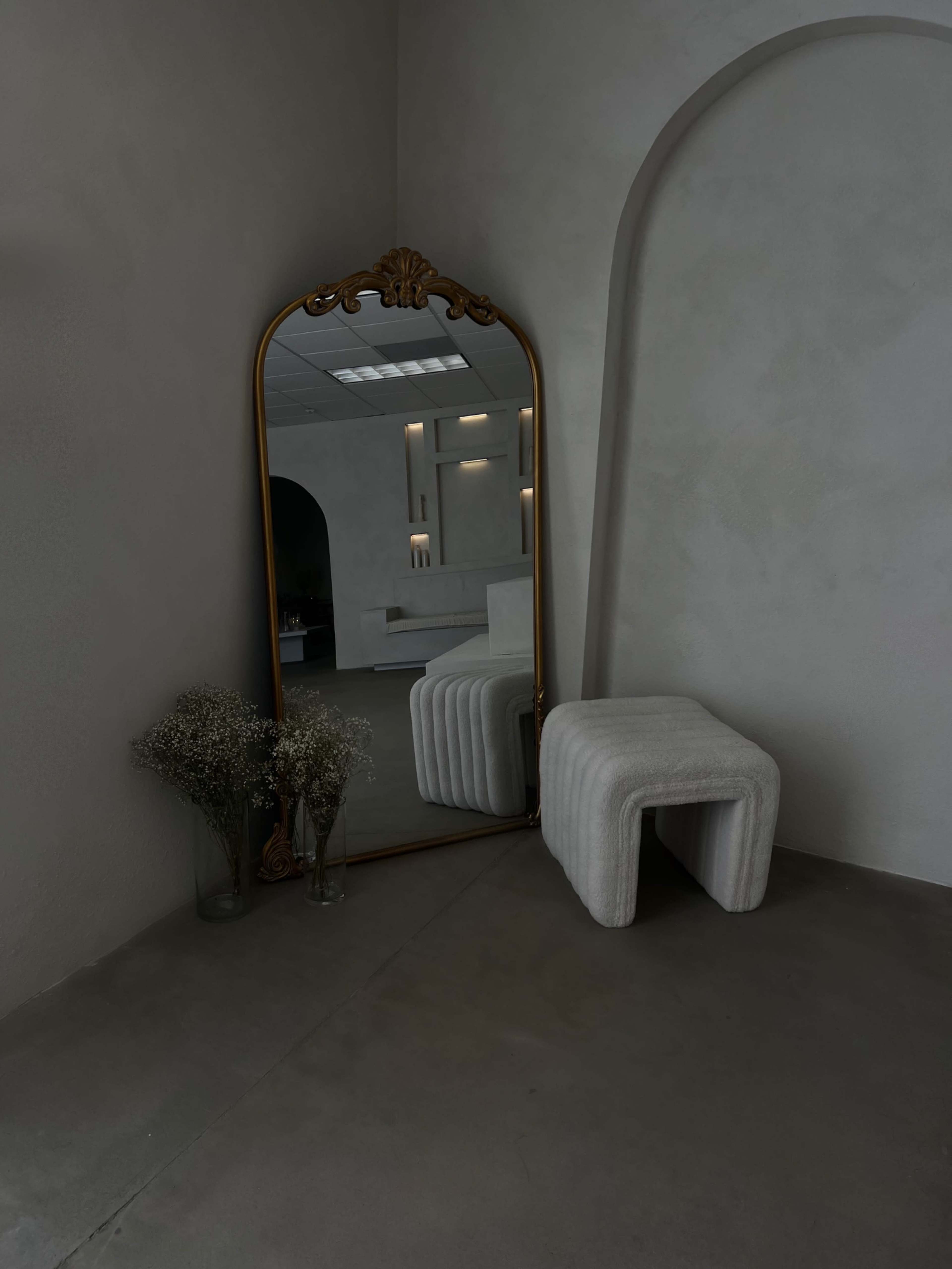 A minimalist room featuring a large ornate mirror, a curved stool, and decorative vases with dried flowers, all set against a light-colored wall.