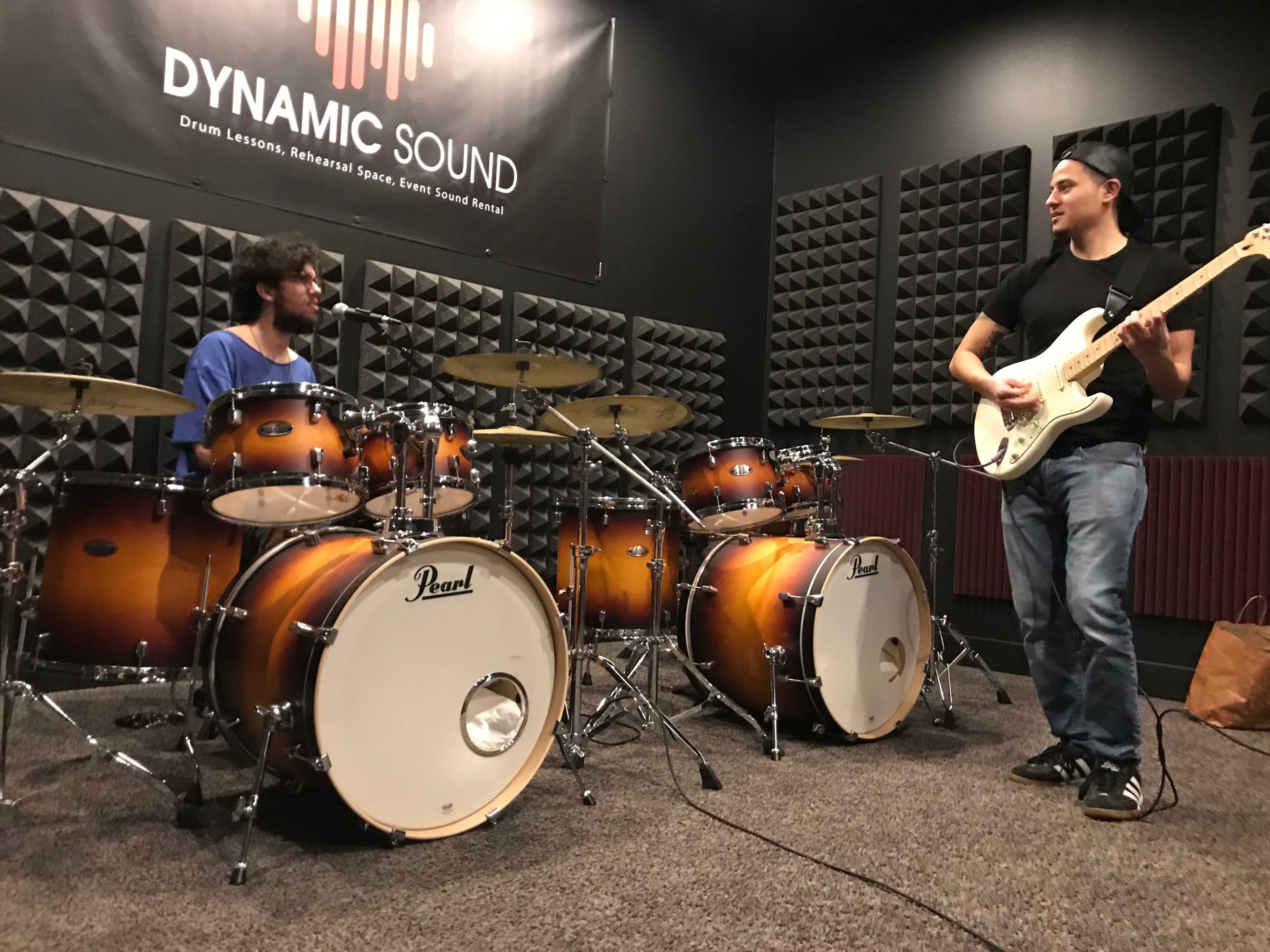 Two musicians are playing music together in a rehearsal space, featuring two drum sets and an electric guitar.