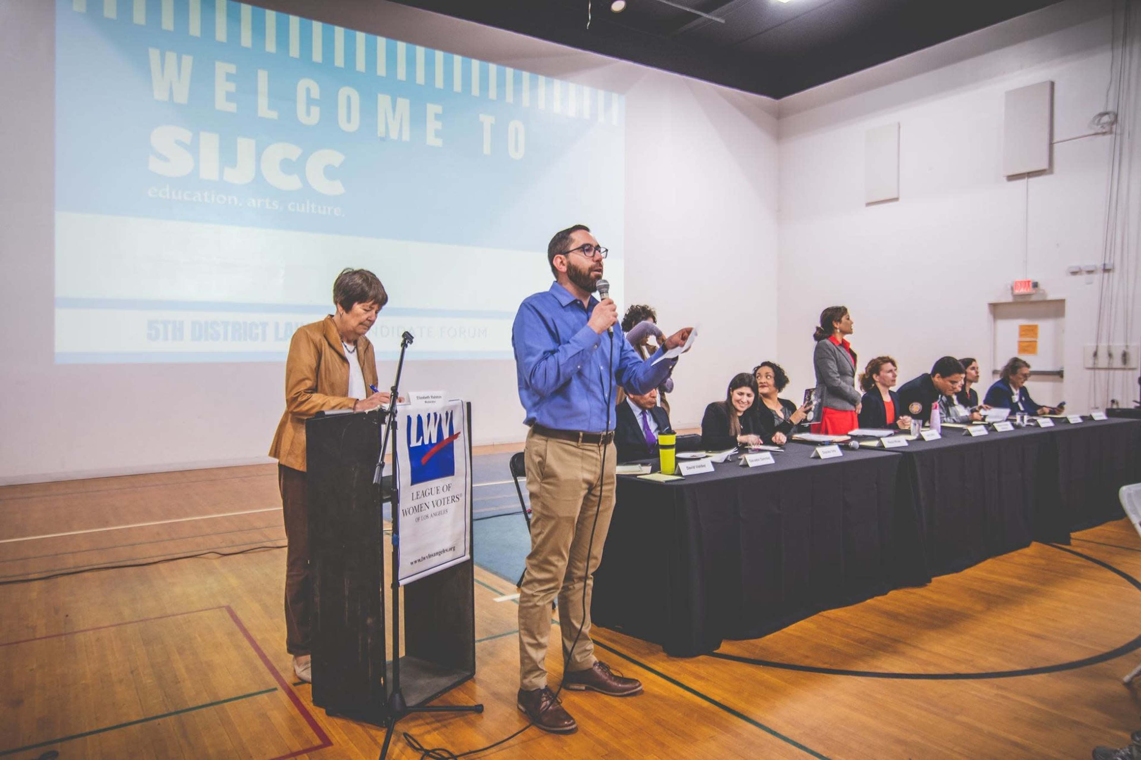 A man at a podium addresses an audience while a woman sits at a table with a group of people, all participating in a public event.