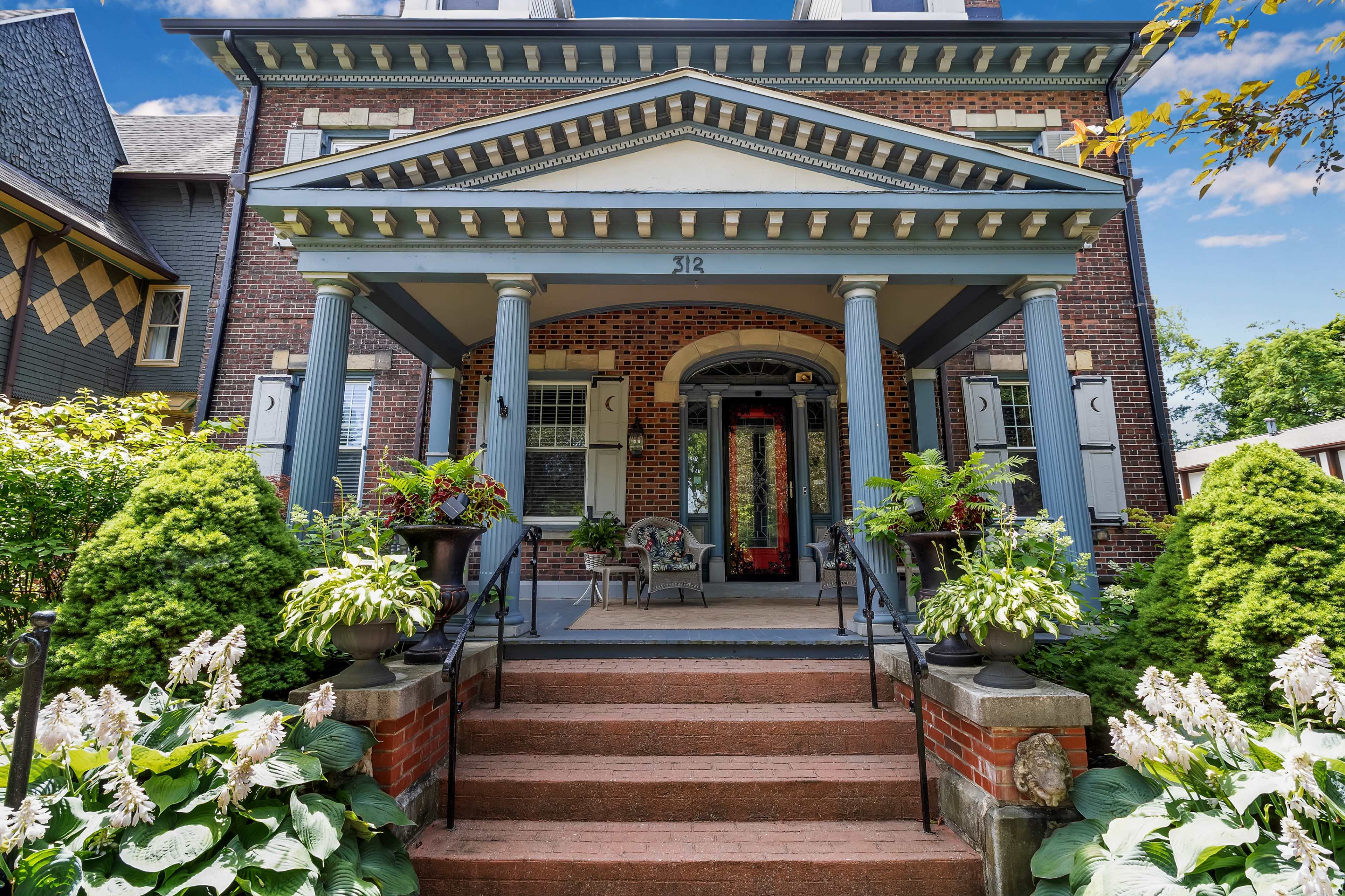 A large brick house features a prominent porch supported by tall blue columns, flanked by well-manicured shrubs and planters.