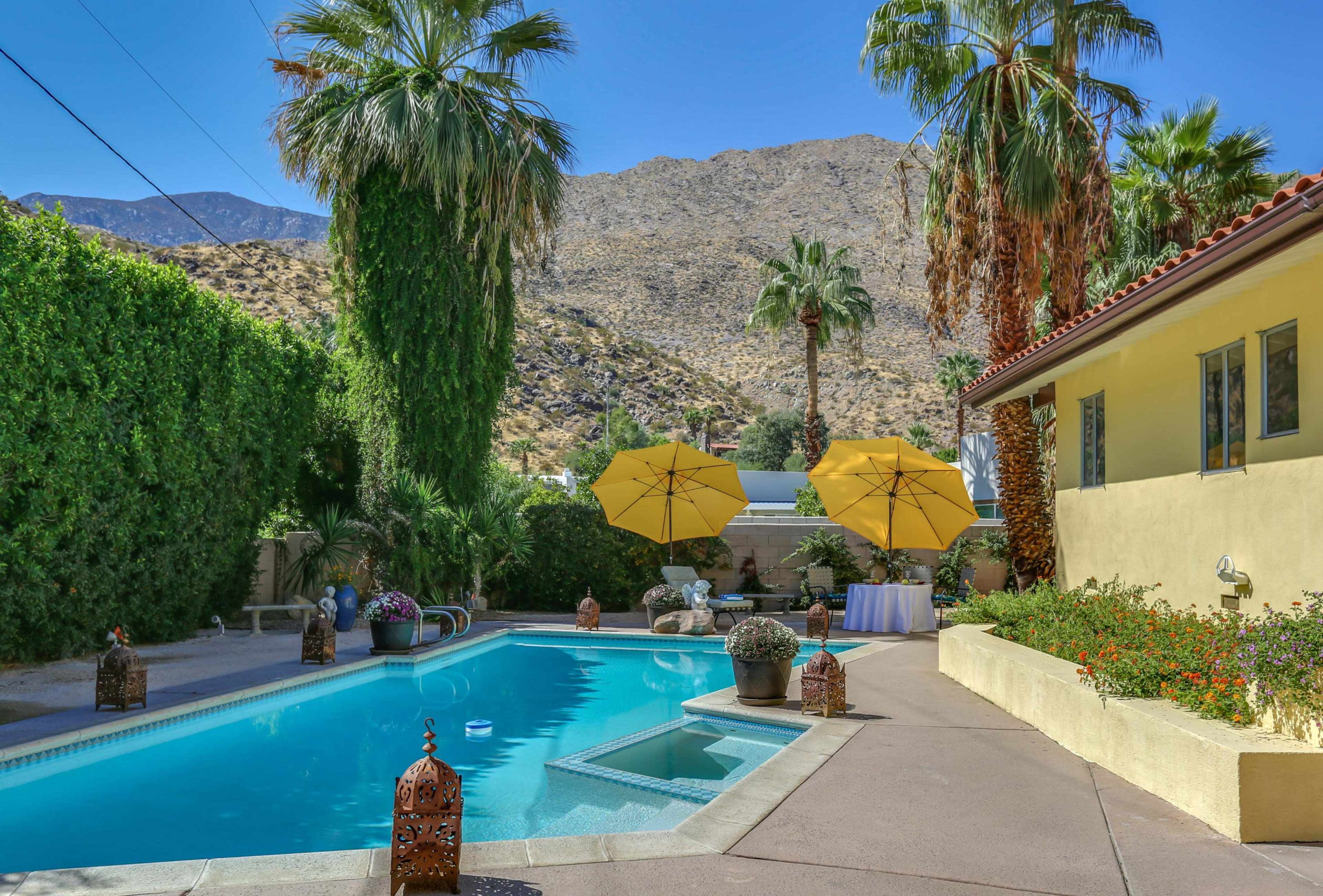A poolside area with yellow umbrellas, surrounded by palm trees and mountain views in the background.