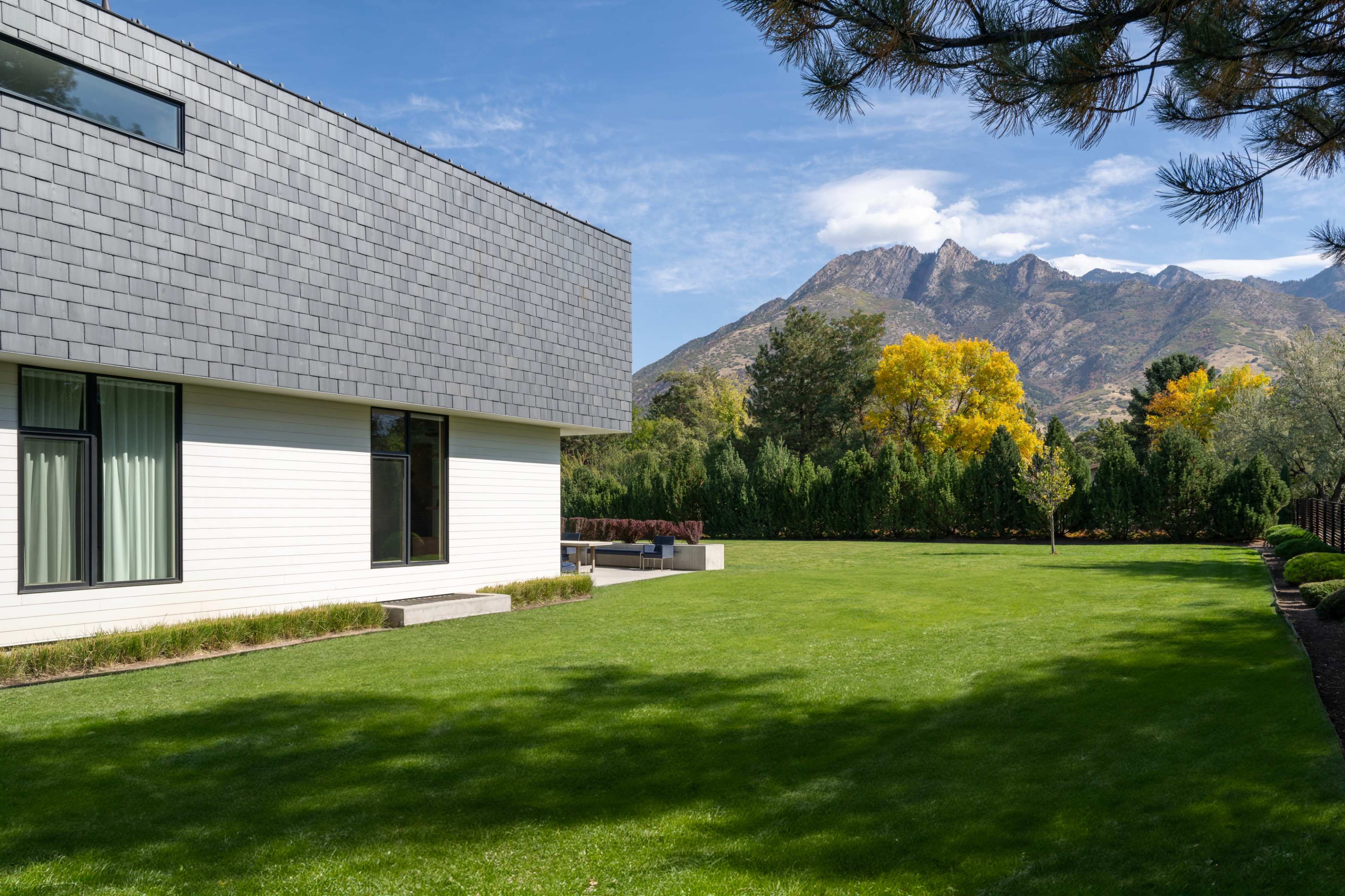 A modern house with a shingled roof sits beside a well-manicured lawn, framed by trees and mountains in the background.