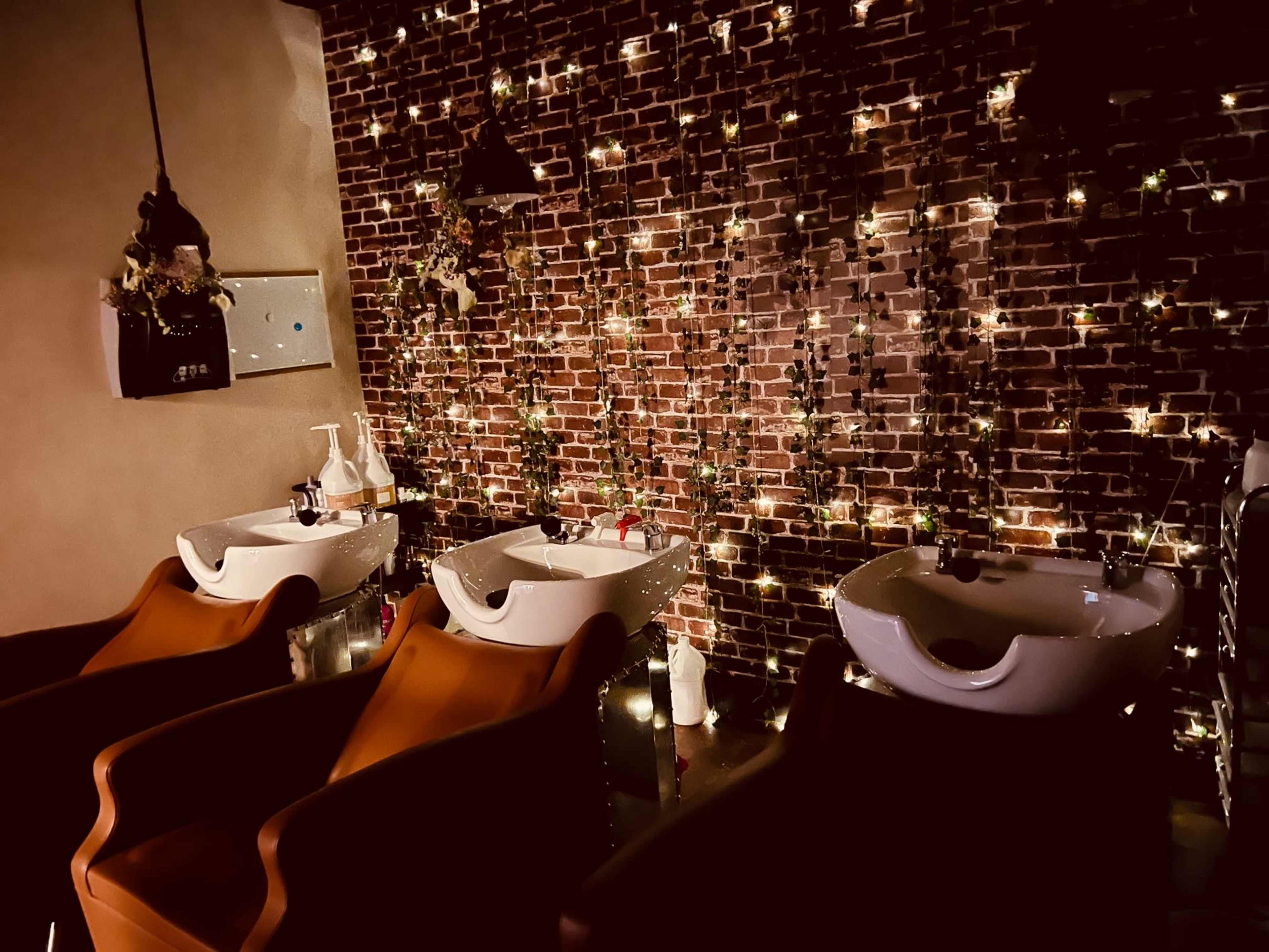 The image shows a salon with three hair washing stations, each featuring a white basin and surrounded by a brick wall decorated with string lights.