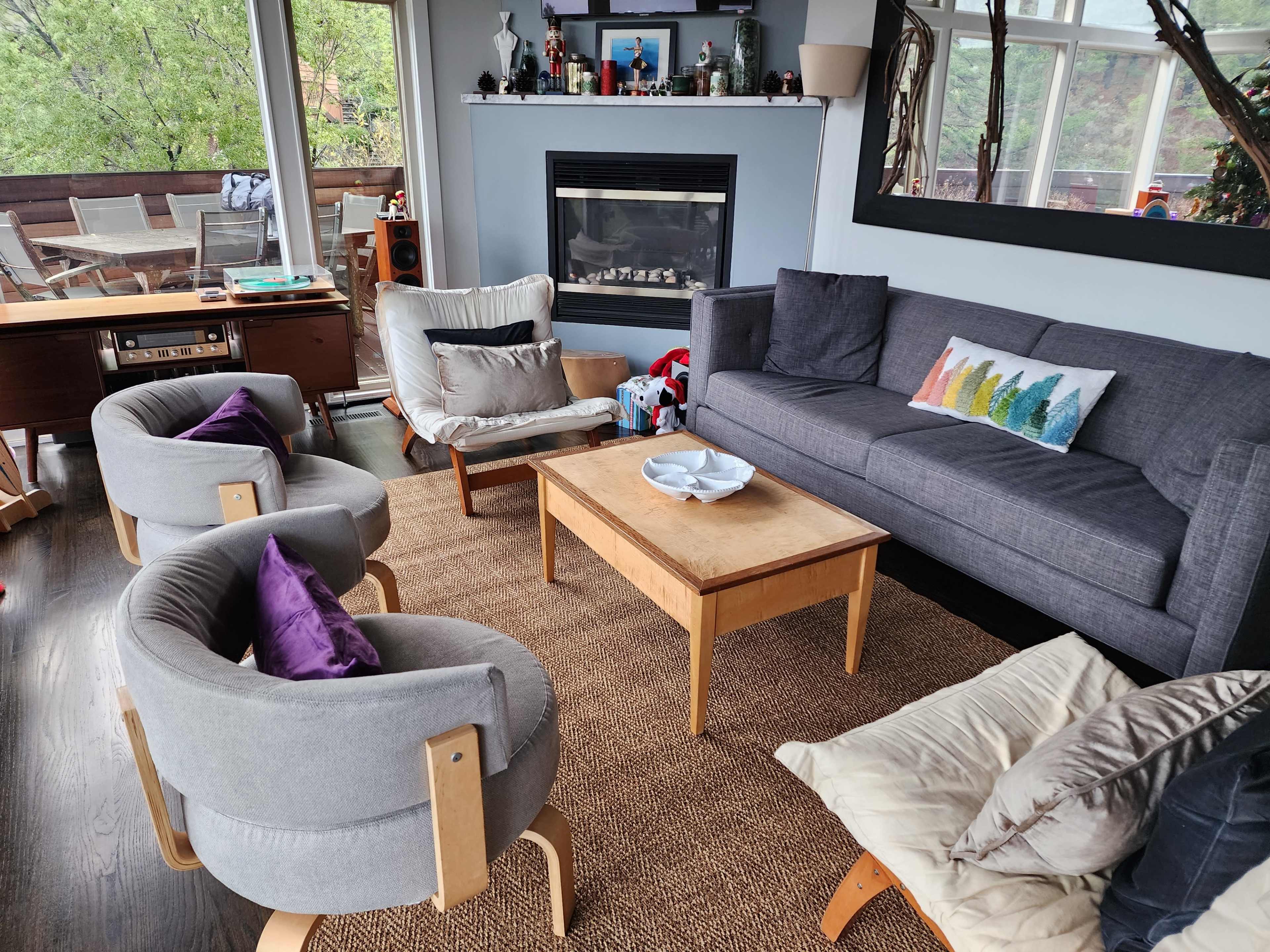 The image shows a living room with a gray sofa, four armchairs, a wooden coffee table, and a fireplace, surrounded by large windows with greenery outside.