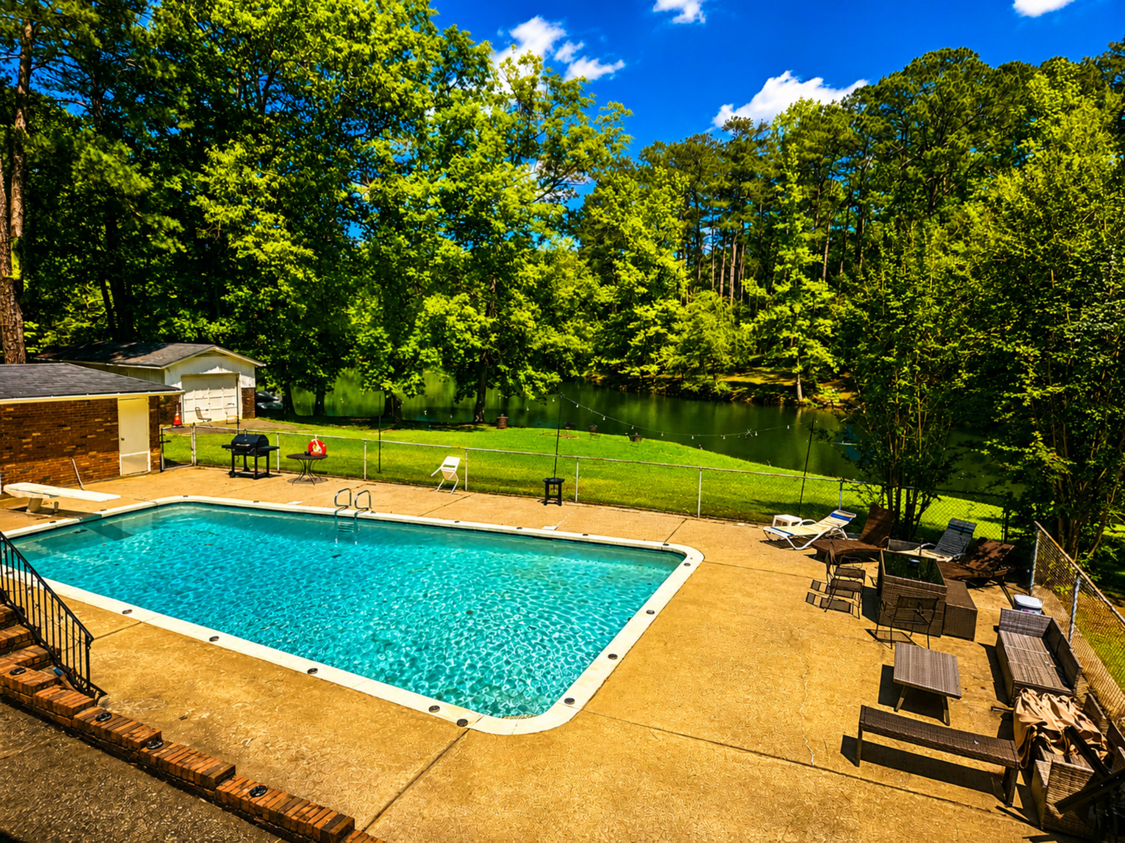 The image shows a clear swimming pool surrounded by a concrete patio, with trees and a grassy area near a body of water in the background.
