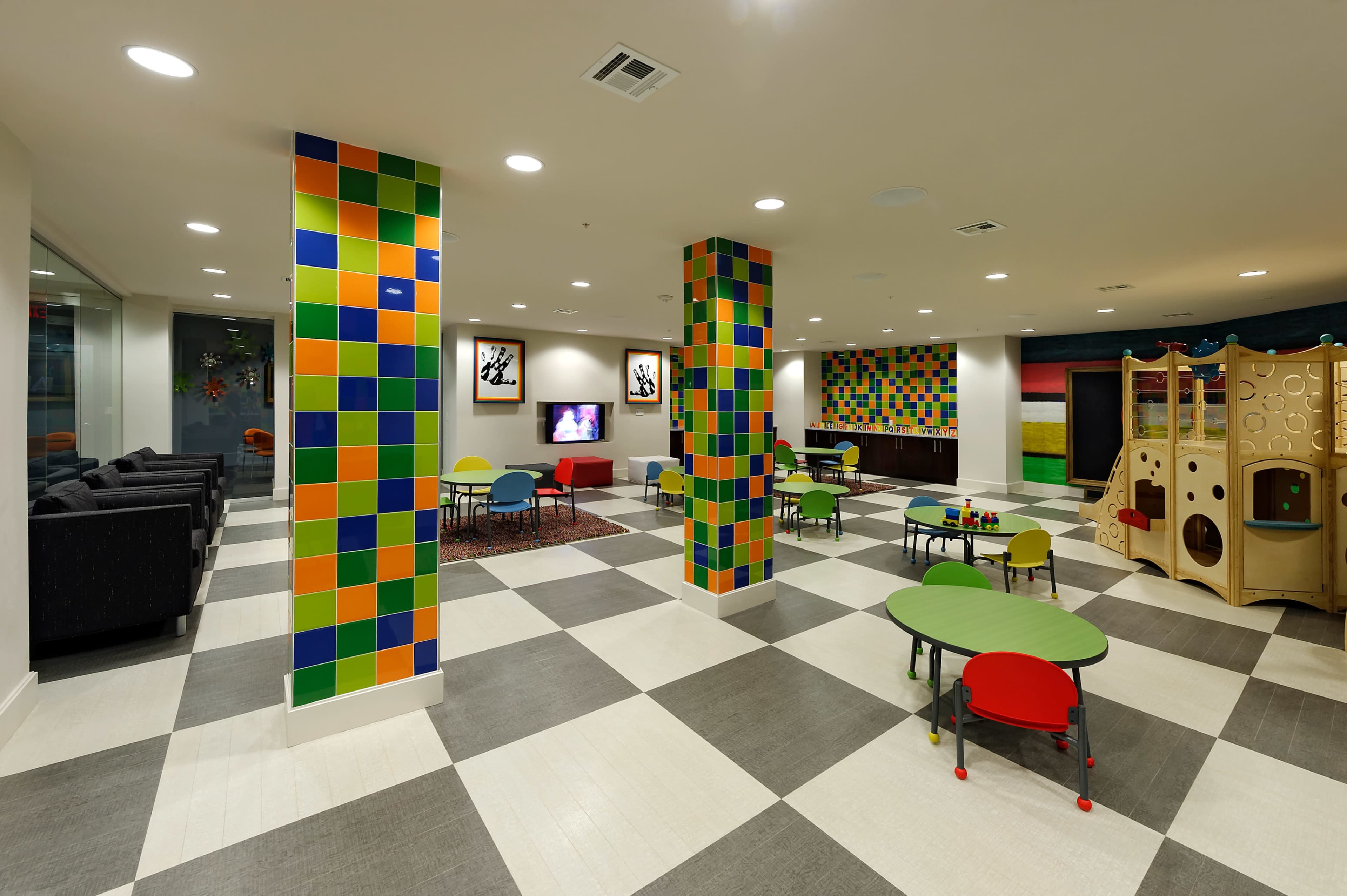 A brightly colored children's playroom with tiled pillars, green tables and chairs, and a play structure.