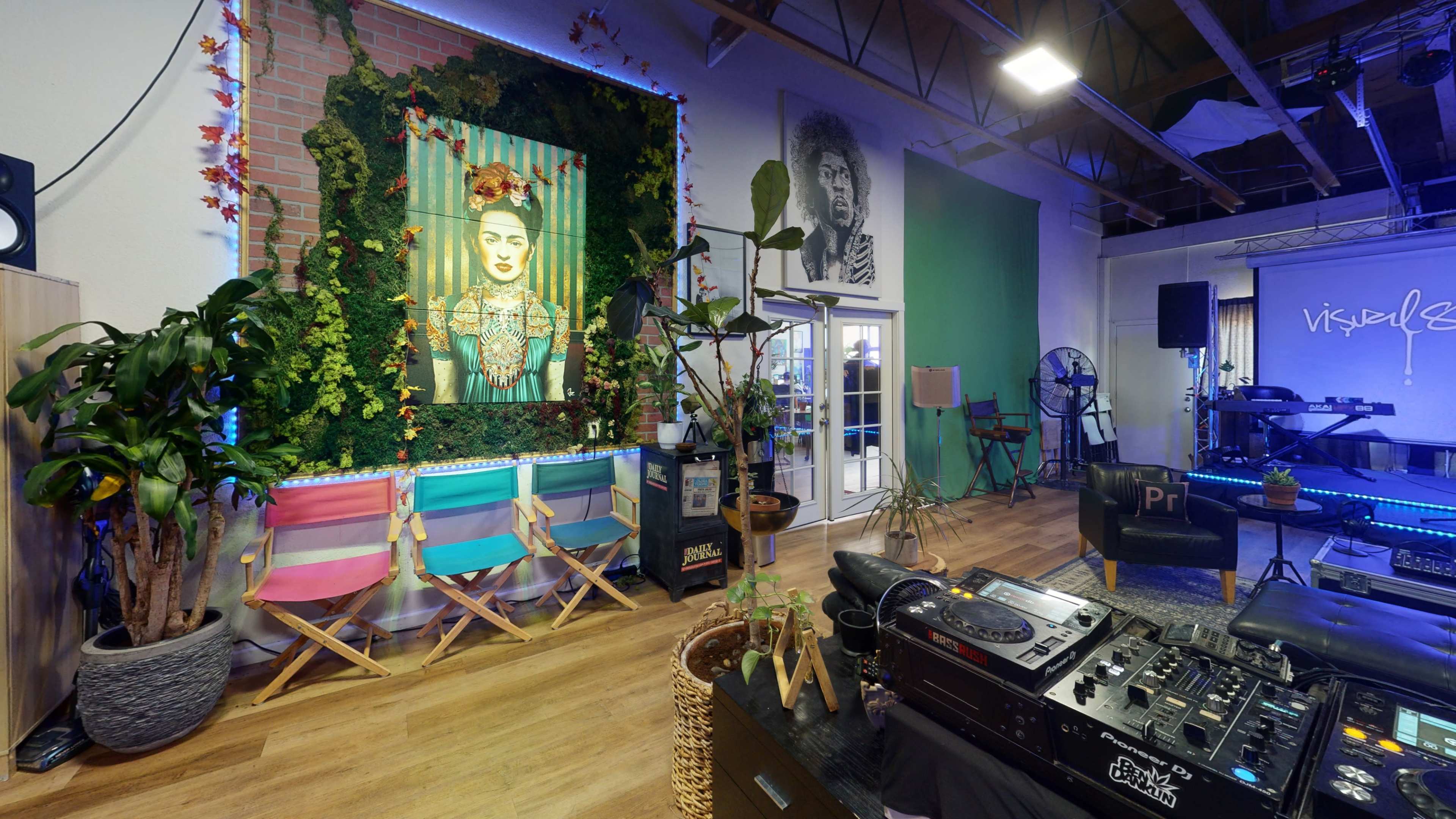 A vibrant creative space featuring a large portrait of Frida Kahlo, a performing stage, colorful chairs, and a DJ setup with plants decoratively arranged throughout.