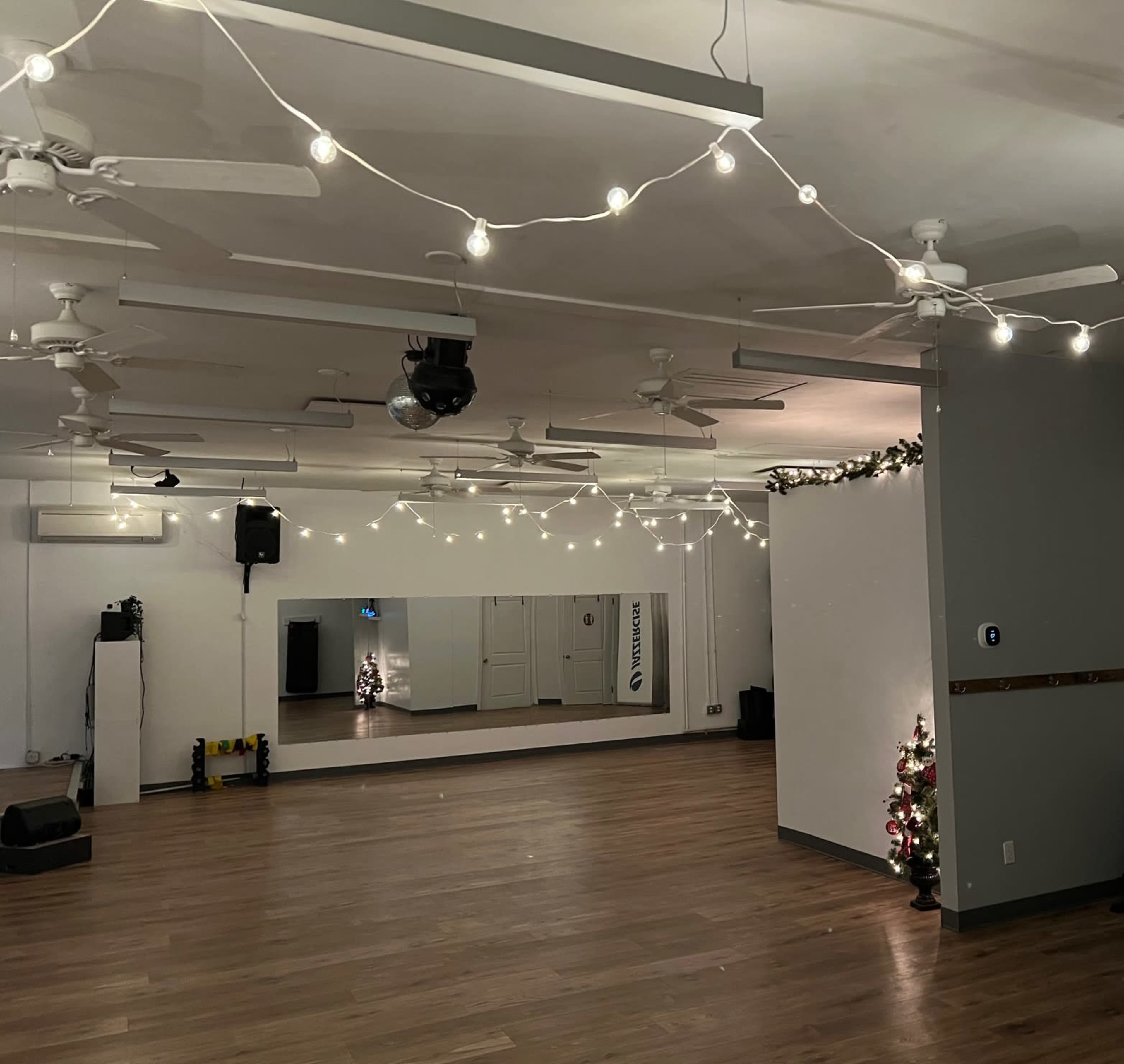 A spacious dance studio with wooden flooring, string lights overhead, a large mirror on one wall, and a small Christmas tree in the corner.