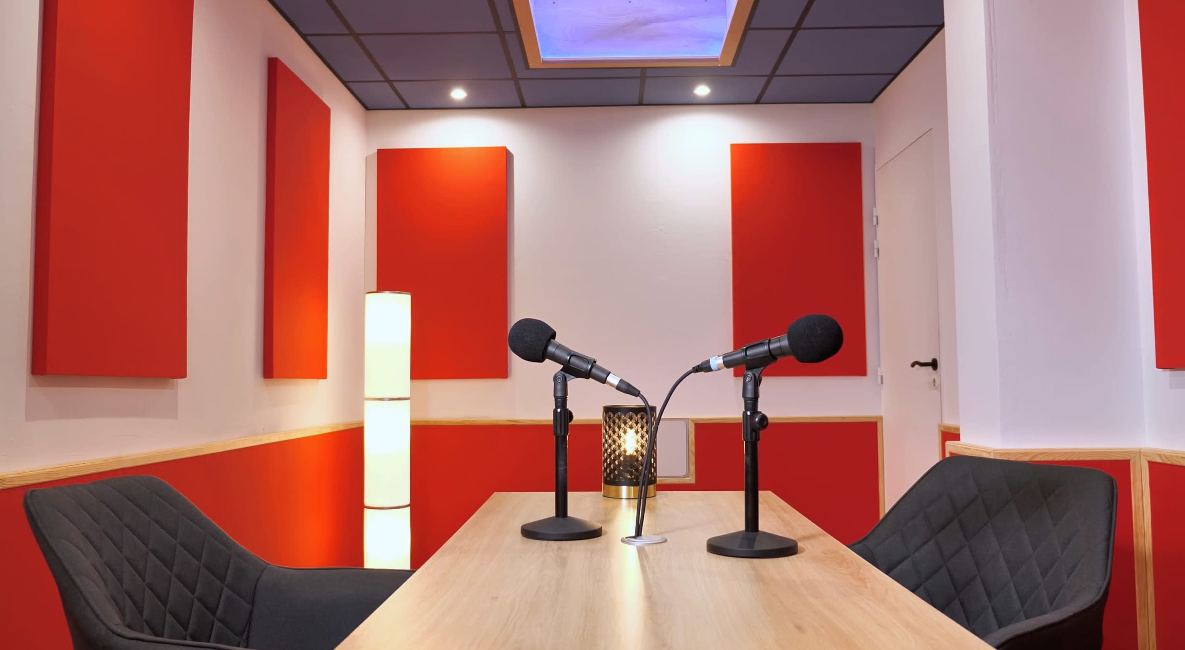 A podcast studio with a wooden table, two microphones, two chairs, and red acoustic panels on the walls.