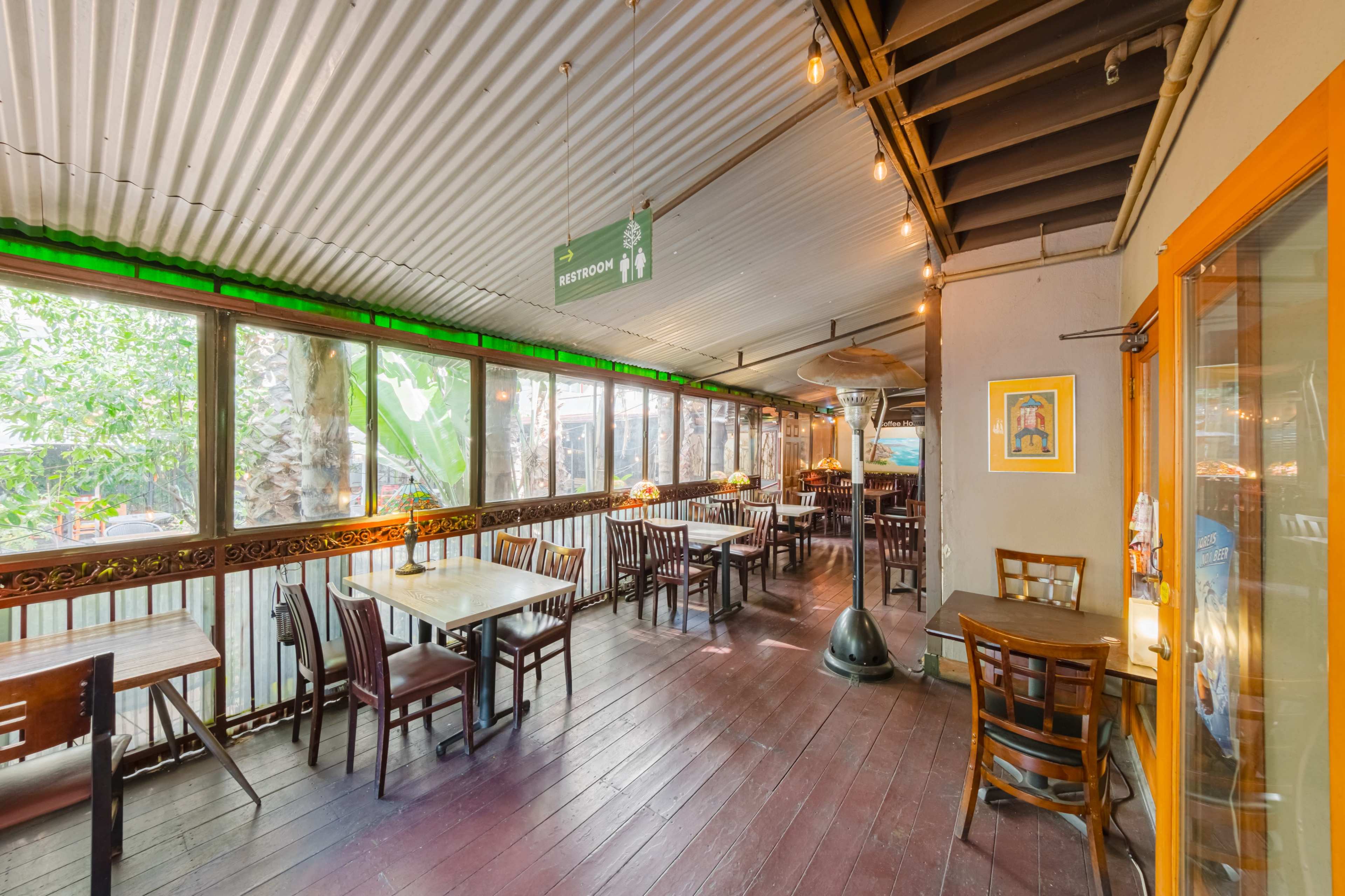 A partially enclosed restaurant with wooden tables and chairs, lit by overhead string lights and surrounded by large windows that provide a view of greenery outside.