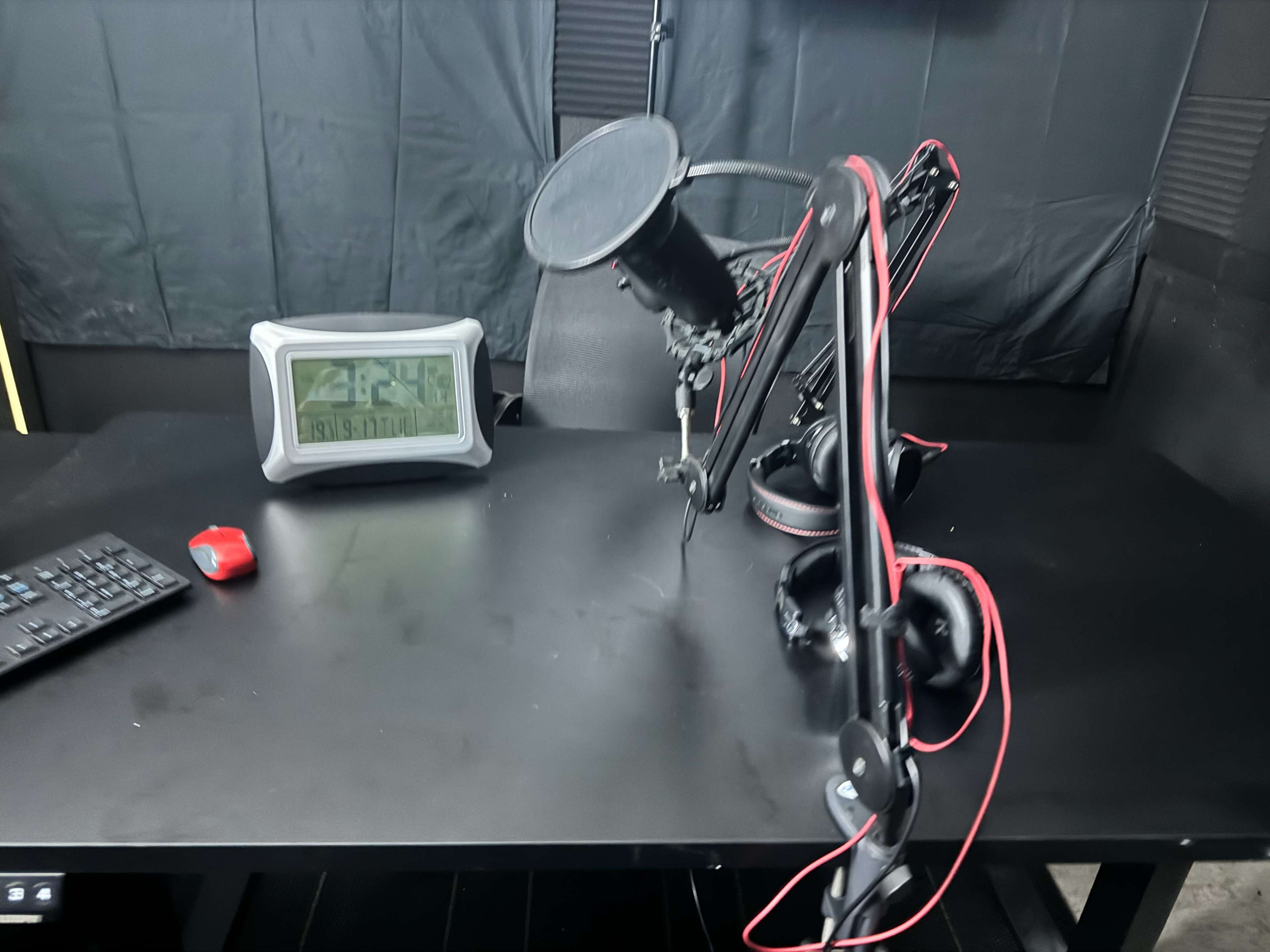 A microphone on an adjustable arm is positioned on a black desk alongside a digital clock and a keyboard.