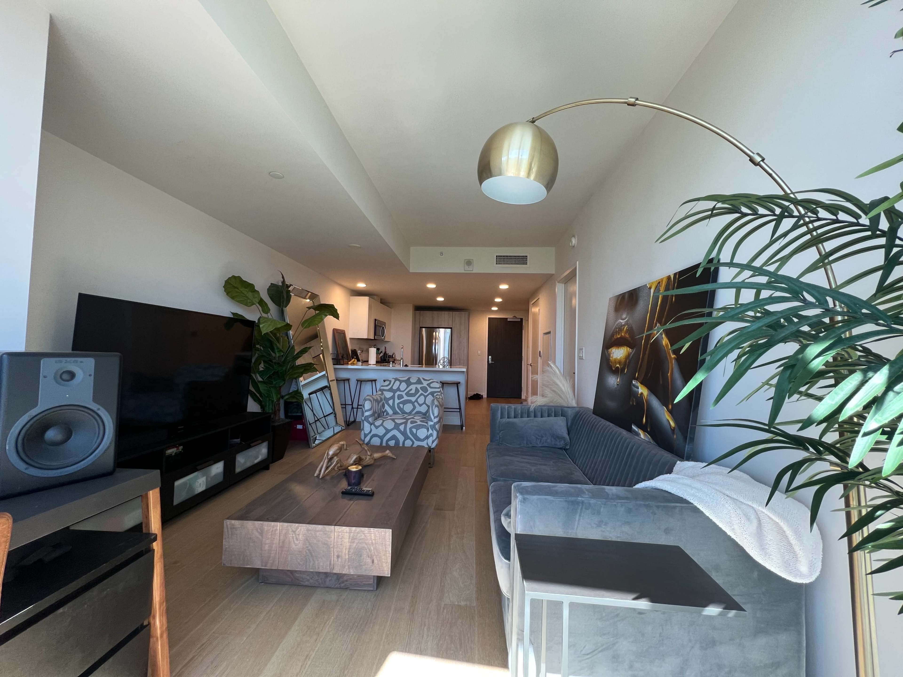 The image shows a modern living room featuring a large television, a sofa, a wooden coffee table, and a kitchen area in the background.