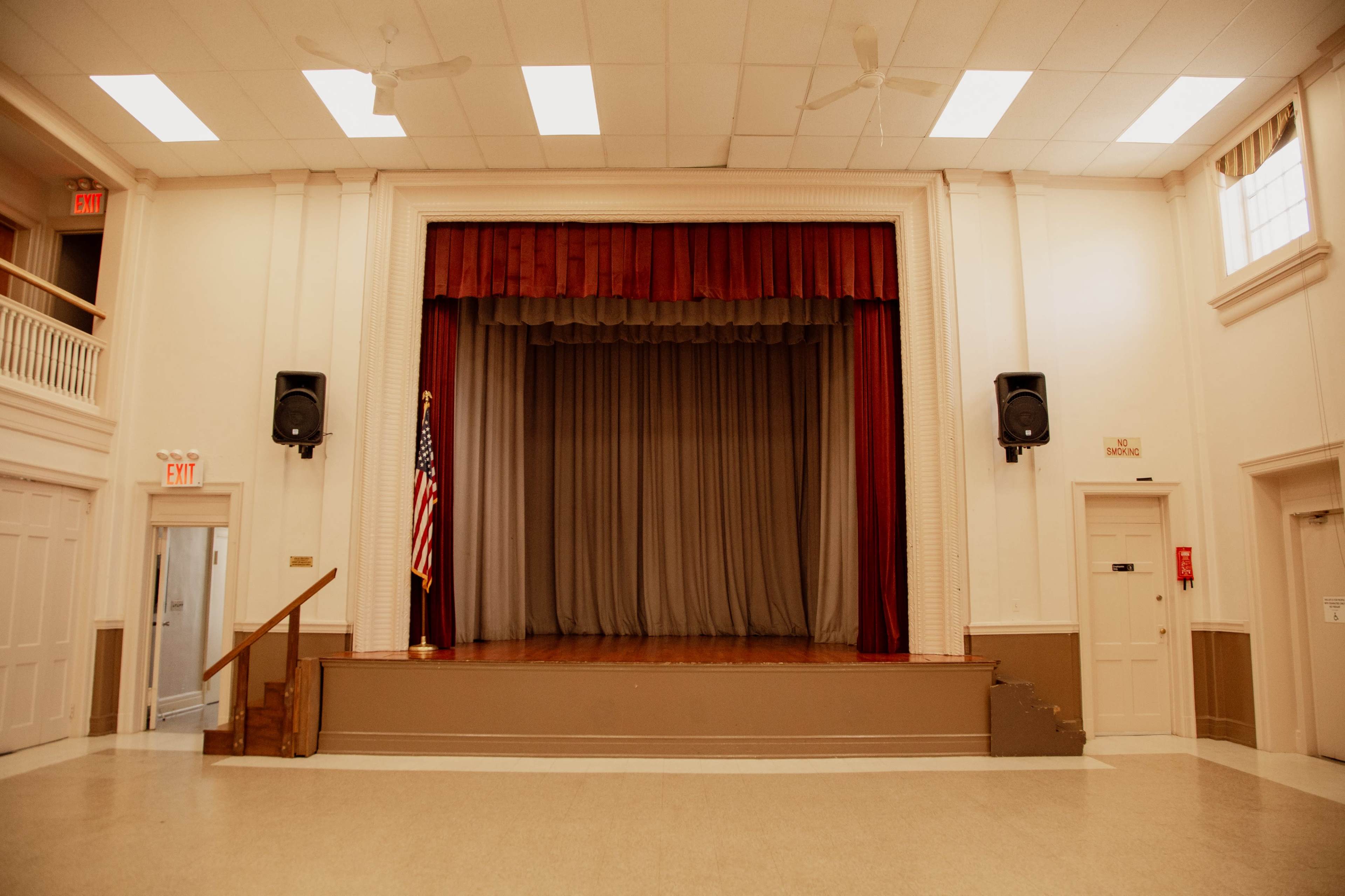 The image shows a vacant stage in a community hall, featuring red curtains, an American flag, and two speakers on either side of the stage.