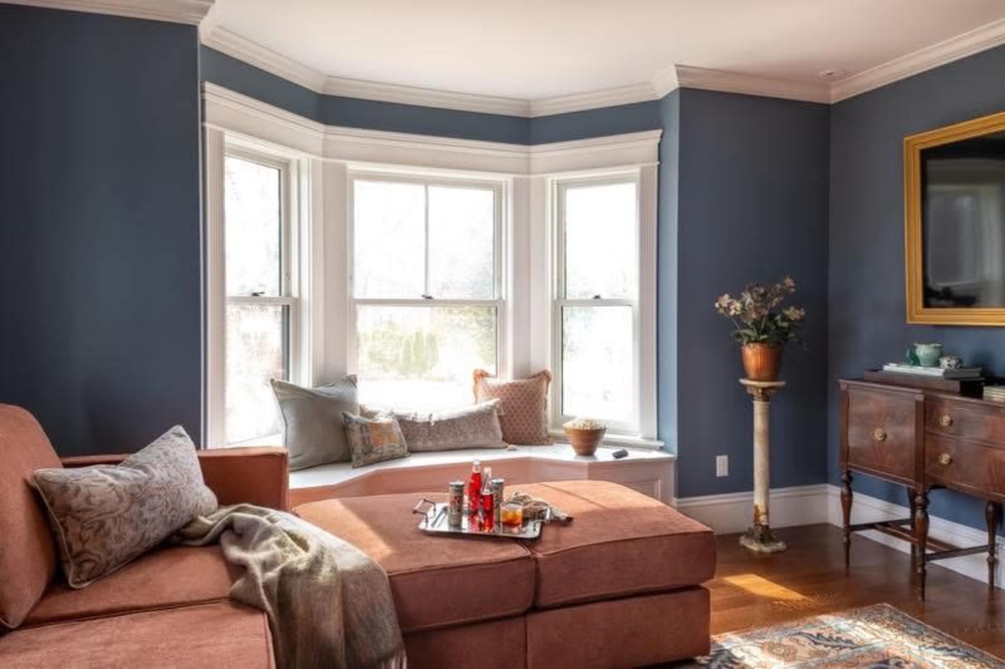 The image features a cozy living room with a large sectional couch, a bay window seat adorned with cushions, and a small table holding snacks and drinks.
