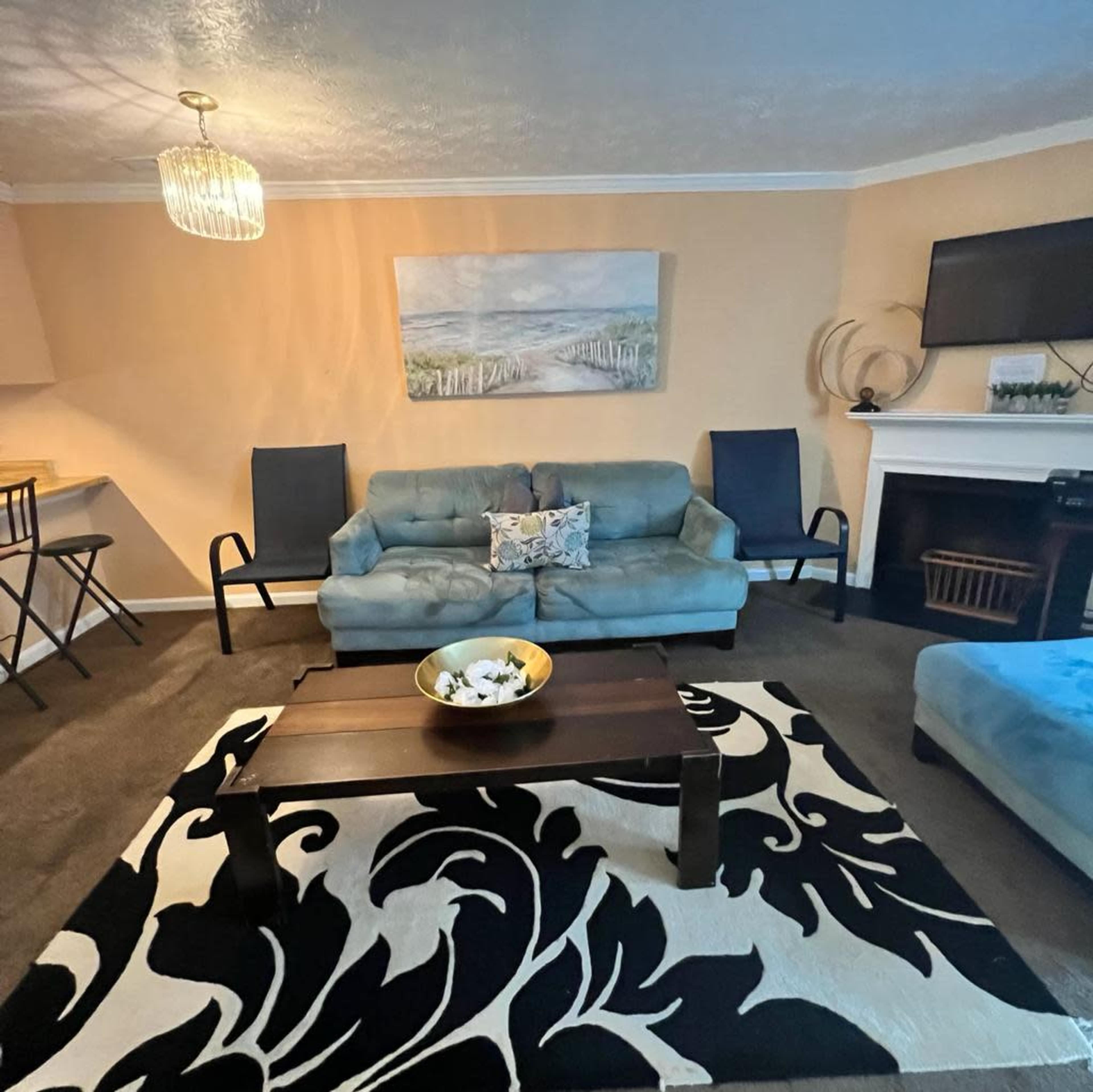 The living room features a blue sofa and two black chairs arranged around a black and white patterned rug, with a wooden coffee table in the center and a large painting on the wall.